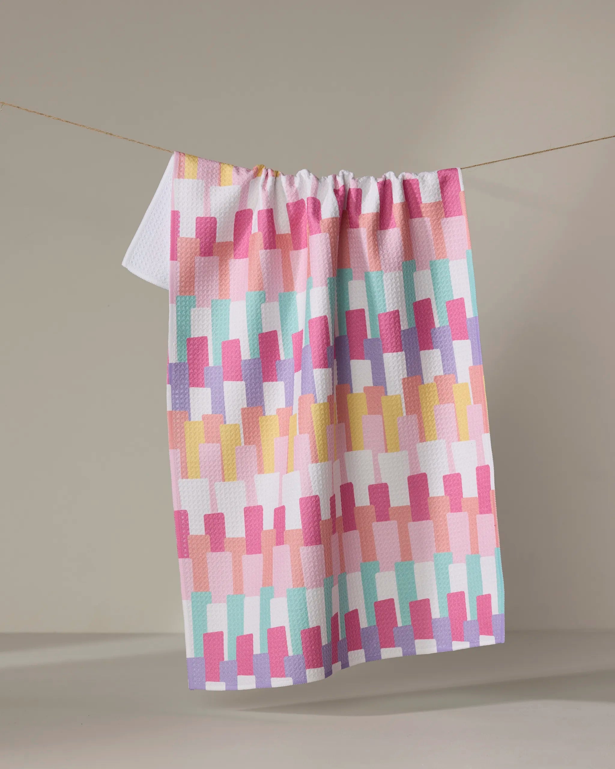 The Keys kitchen towel by Rebecca Bobko features a geometric pattern of overlapping rectangles in pastel pink, yellow, purple, and turquoise—made from recycled materials—hanging on a line against a neutral background.