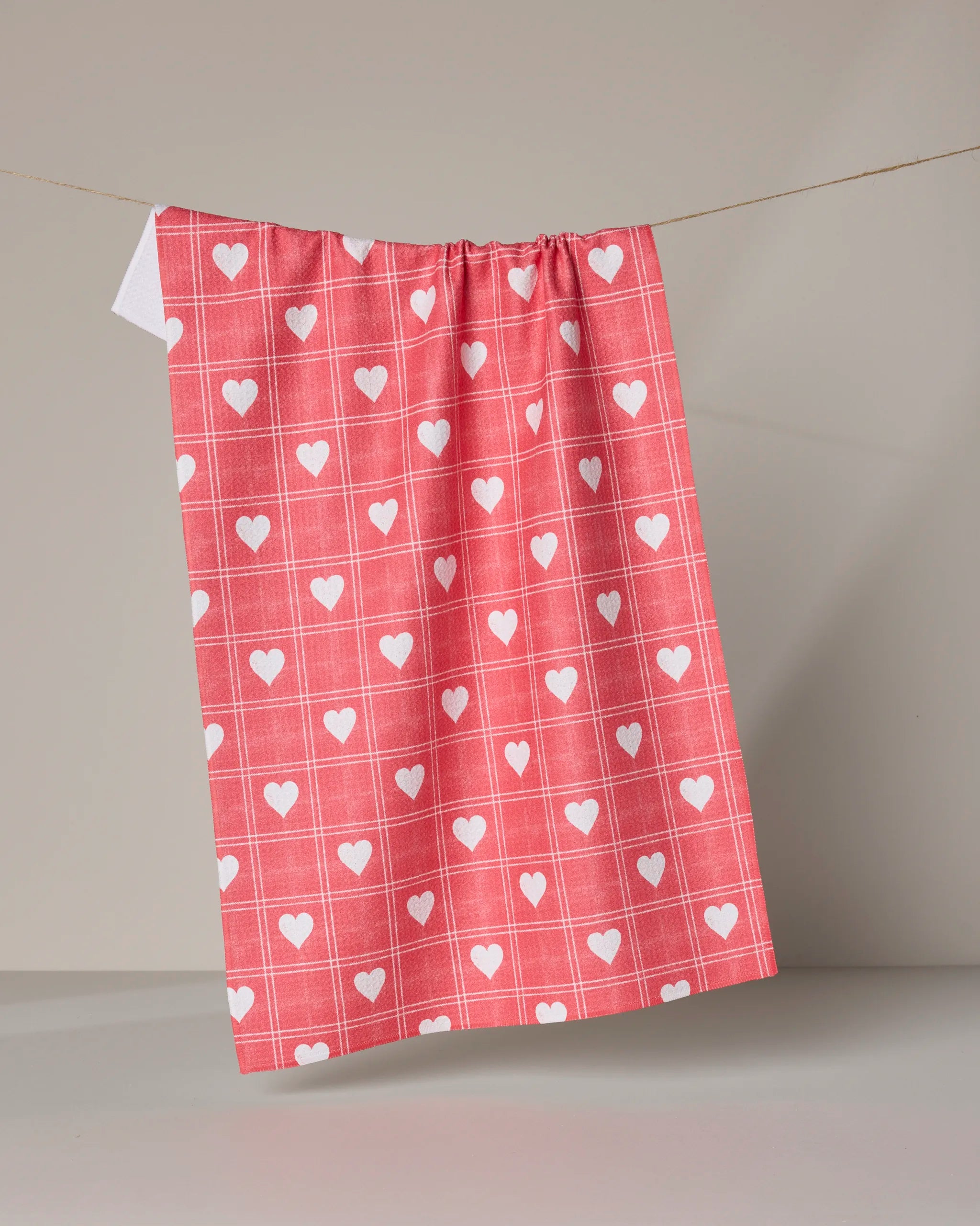 The Geometry Kind Spirit Red tea towel, featuring a red and white checkered design with white hearts, is made from ultra-absorbent recycled materials and hangs on a string against a neutral background.