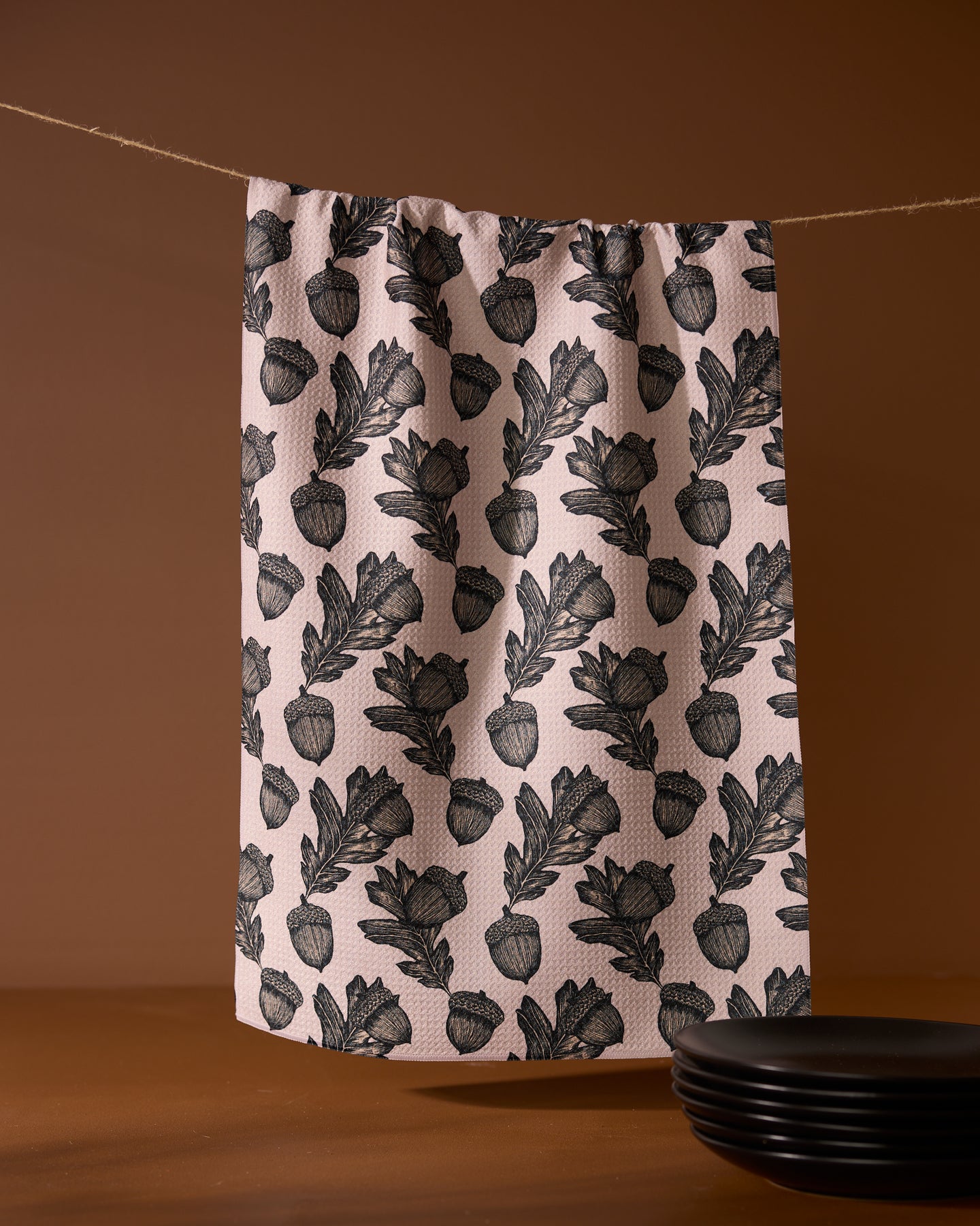 The Kindred kitchen towel by Geometry, featuring a black botanical pattern on pink recycled fabric, hangs on a clothesline against a brown background, with black plates stacked in the lower right corner.
