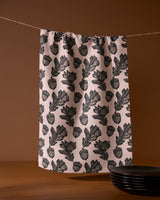 The Kindred kitchen towel by Geometry, featuring a black botanical pattern on pink recycled fabric, hangs on a clothesline against a brown background, with black plates stacked in the lower right corner.