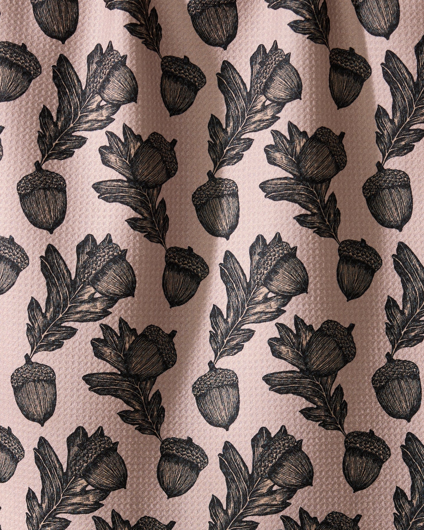 Geometry’s Kindred dish towel features an illustrated pattern of black acorns and oak leaves, evenly spaced on a textured light pink background. Ultra-absorbent fabric forms soft folds with subtle shadows and highlights.