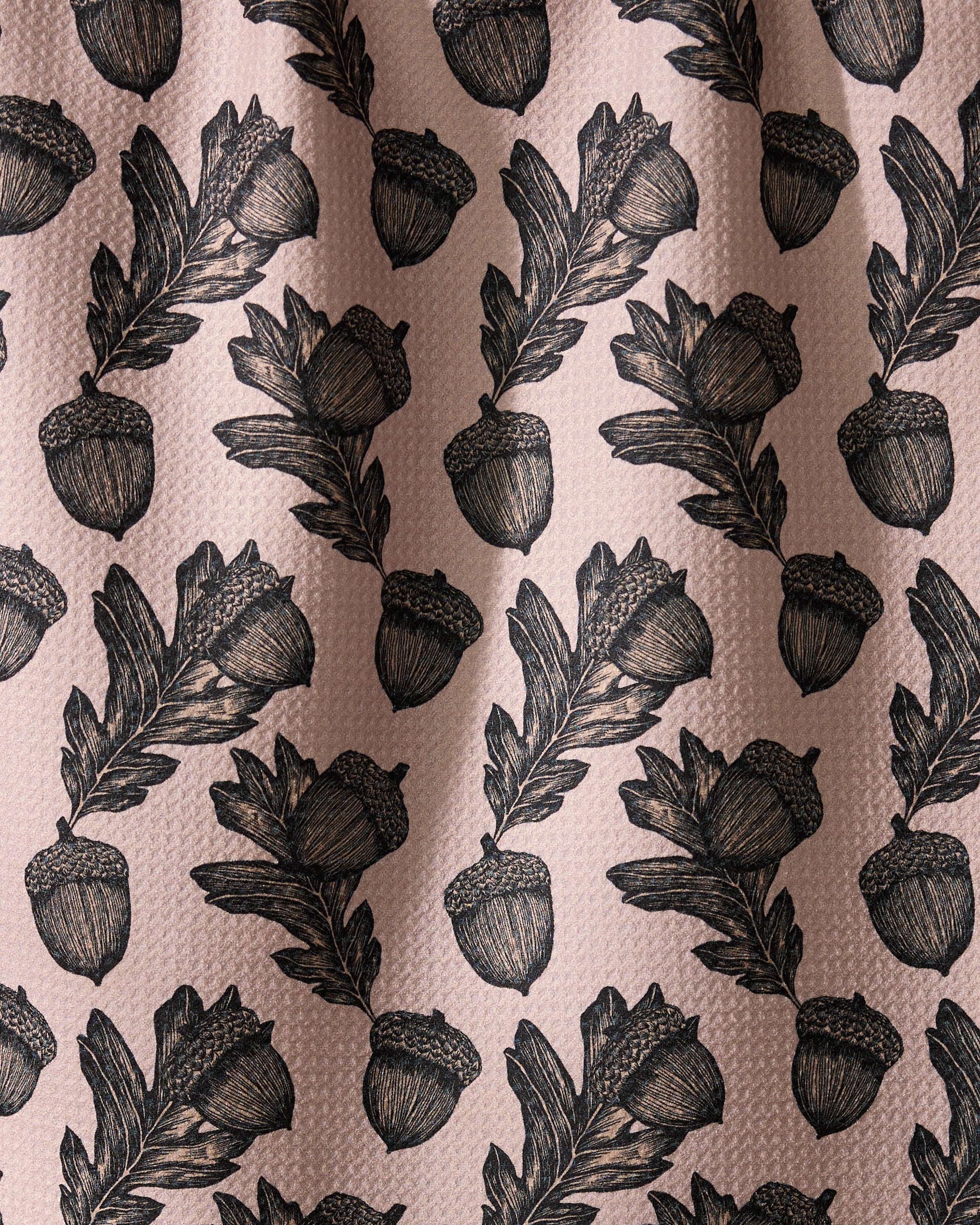 Geometry’s Kindred dish towel features an illustrated pattern of black acorns and oak leaves, evenly spaced on a textured light pink background. Ultra-absorbent fabric forms soft folds with subtle shadows and highlights.