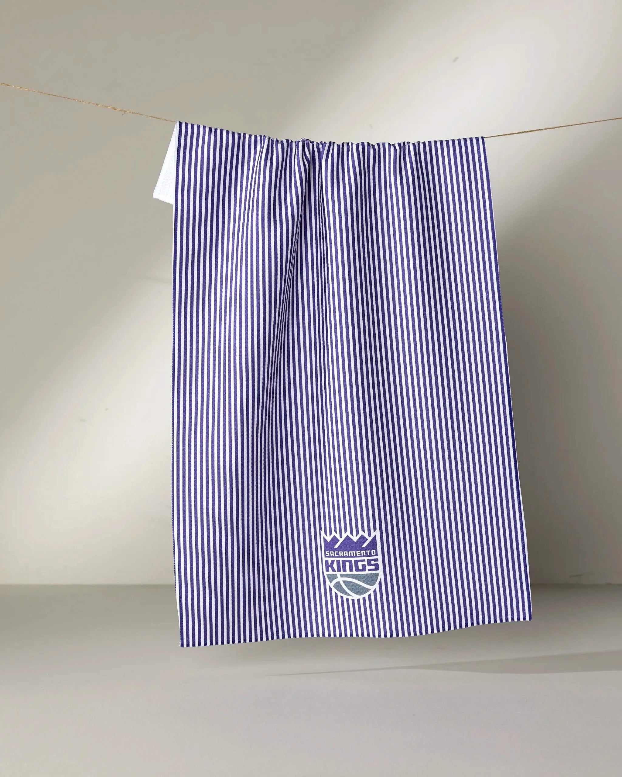 A white and purple vertical striped Kings Classic tea towel by NBALAB x Geometry hangs from a line, featuring the Sacramento Kings logo near the bottom center.