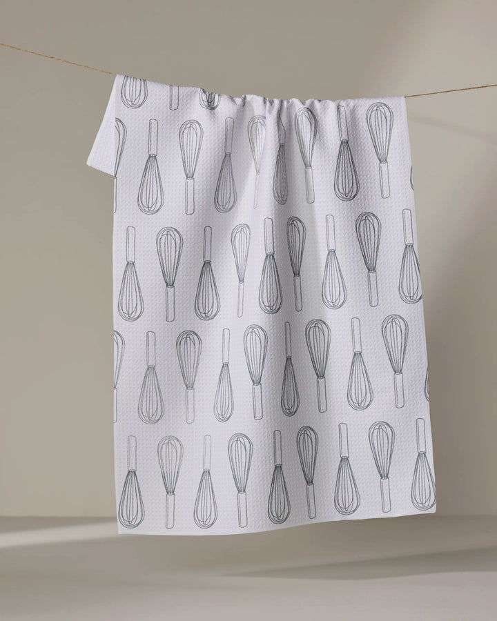 A “Kitchen Whisks” tea towel by Rosa Kichline, made from quick-drying recycled materials, hangs on a line. It features a repeating black wire whisk pattern on white and is set against a softly lit neutral background.