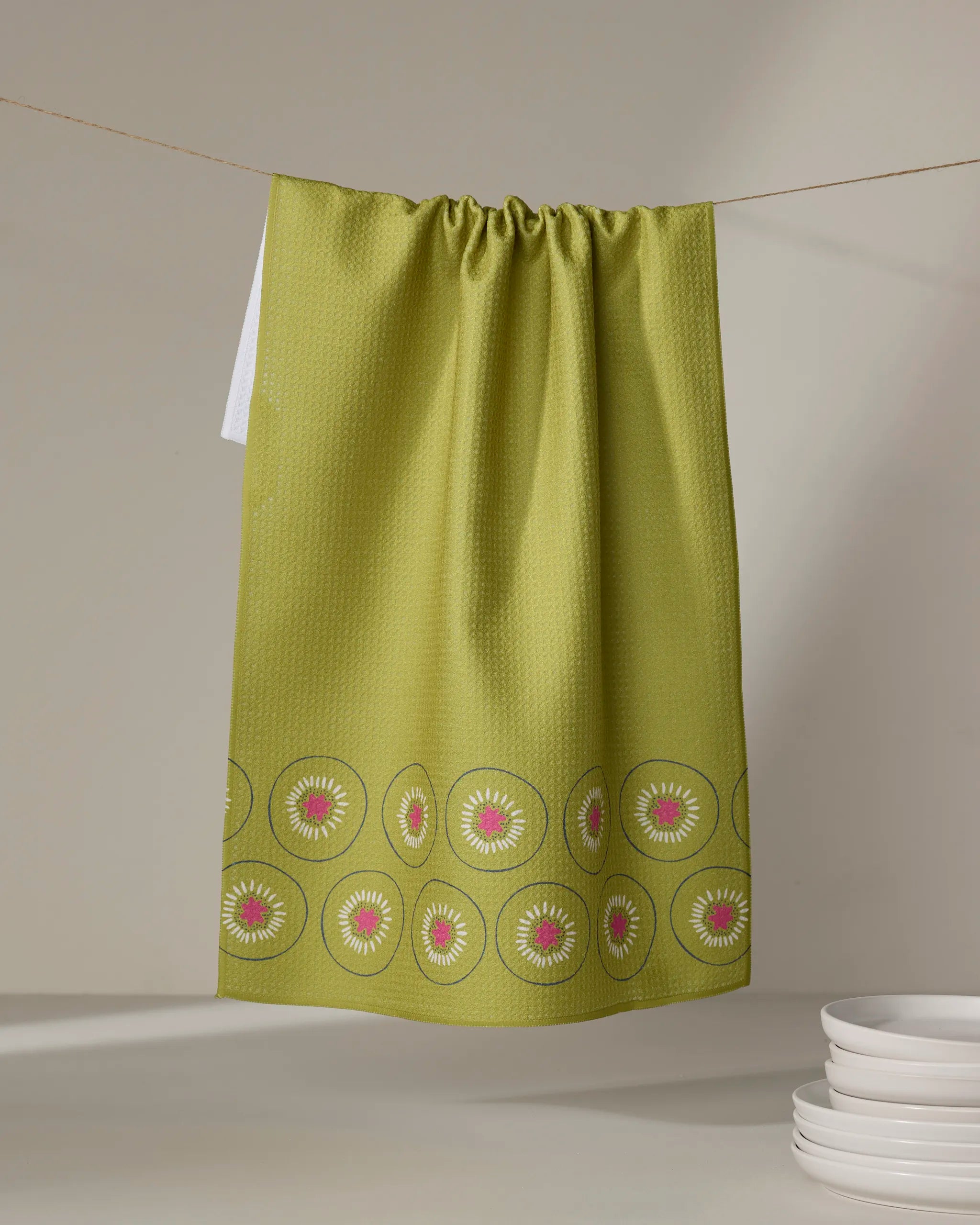 The Lizzie Clark "Kiwi Kraze" kitchen tea towel, featuring a border of stylized kiwi slices, hangs on a string above stacked white plates. The soft neutral background highlights the towel’s vibrant green design.