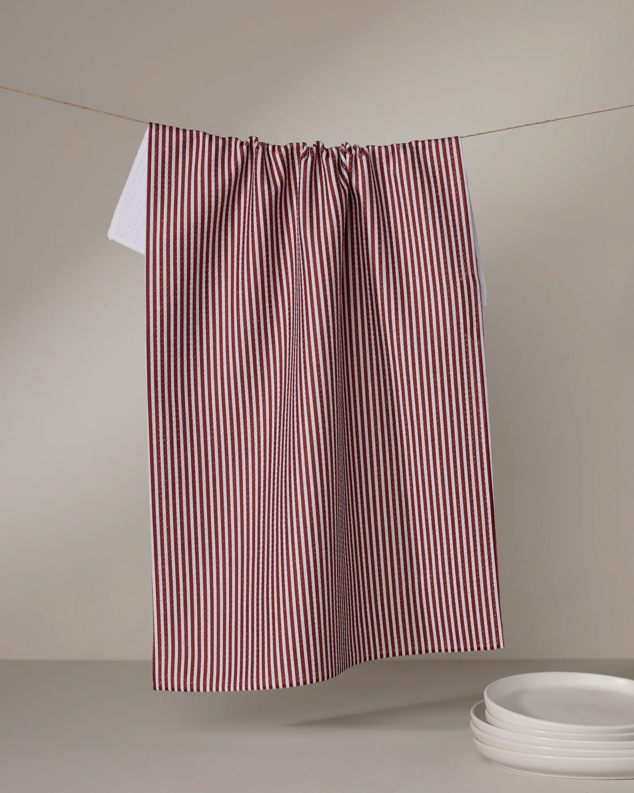 The Geometry Kringle tea towel, featuring red and white vertical stripes and made from recycled materials, hangs on a string above neatly stacked white plates against a plain background.
