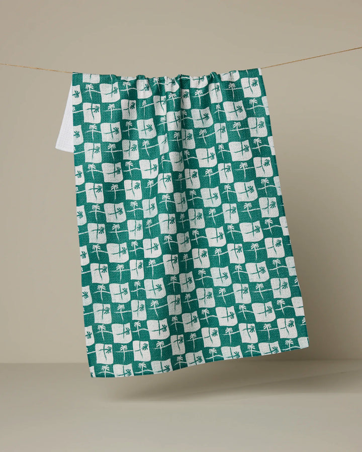 The La Brisa kitchen towel by Geometry, made from recycled materials, displays a green and white checkered leaf and flower pattern and hangs over a string against a plain beige background.