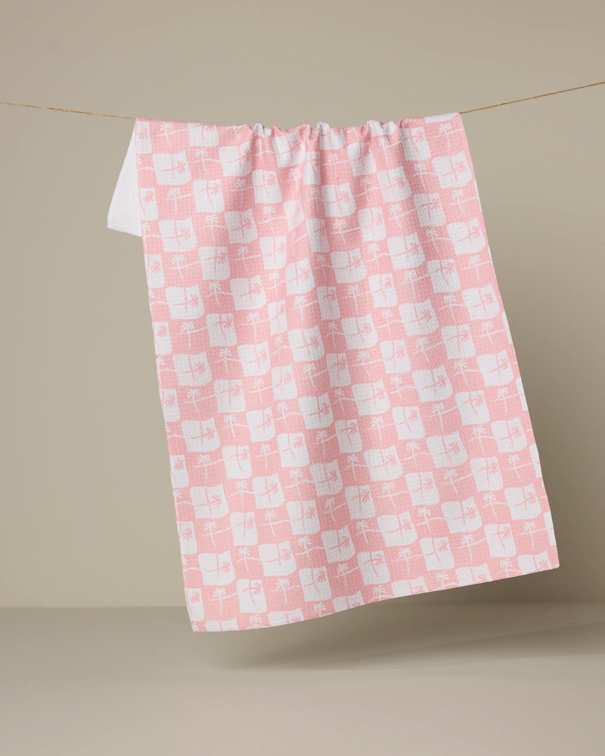 The La Brisa tea towel by Geometry, made from recycled materials, hangs on a thin string. It features a pink and white checkered geometric design with abstract shapes against a plain beige background.