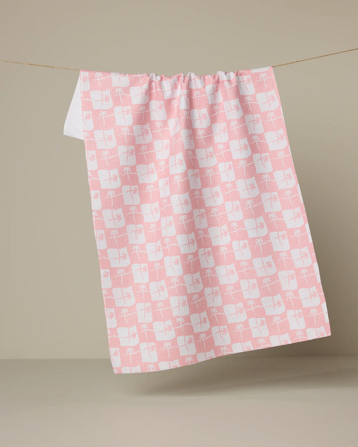 The La Brisa tea towel by Geometry, made from recycled materials, hangs on a thin string. It features a pink and white checkered geometric design with abstract shapes against a plain beige background.