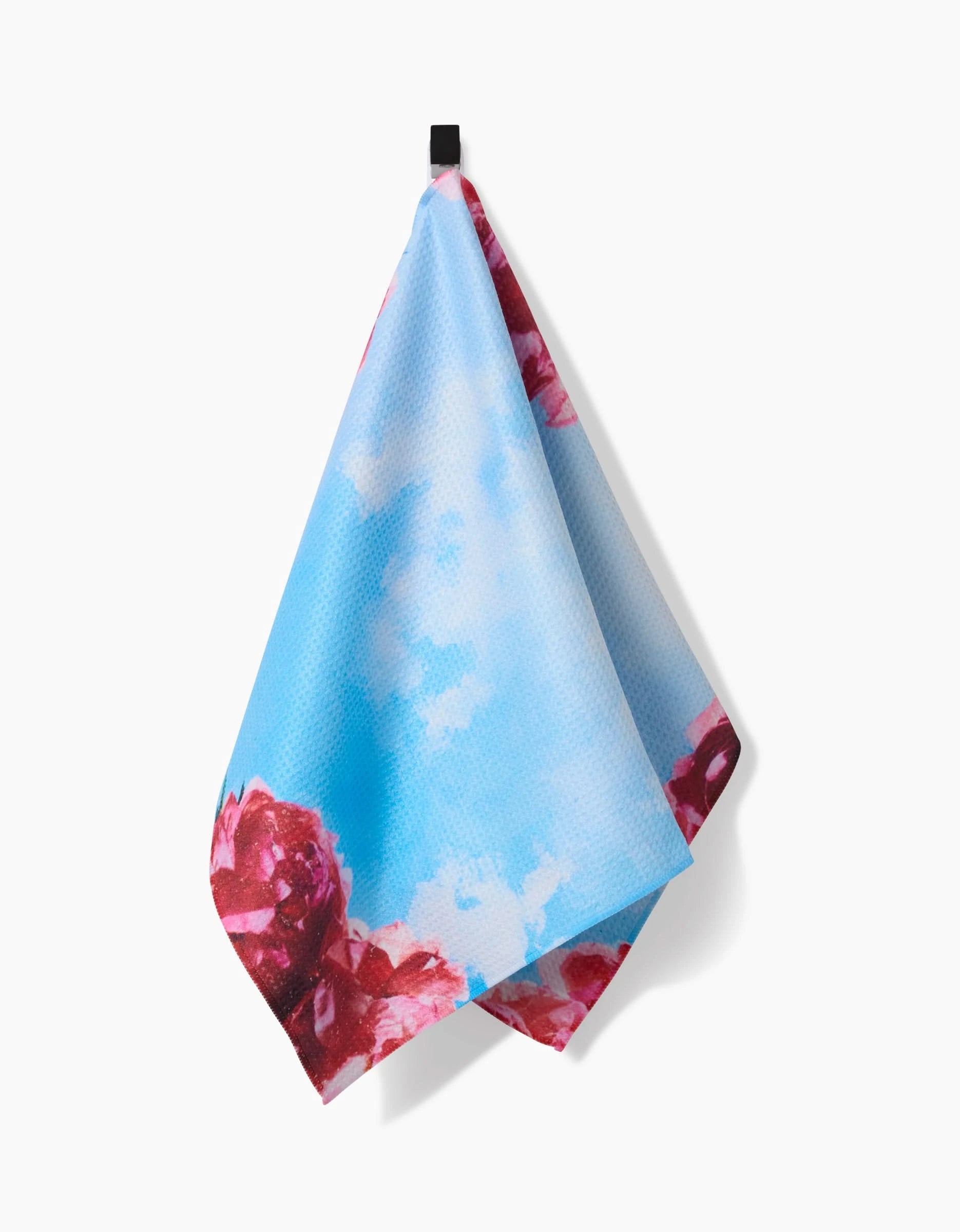 The La Floraison tea towel by Stefanie Bales features a blue sky, white clouds, and red and pink flowers at the base. Hanging from a black loop on a white background, it’s made from recycled materials for eco-friendly appeal.