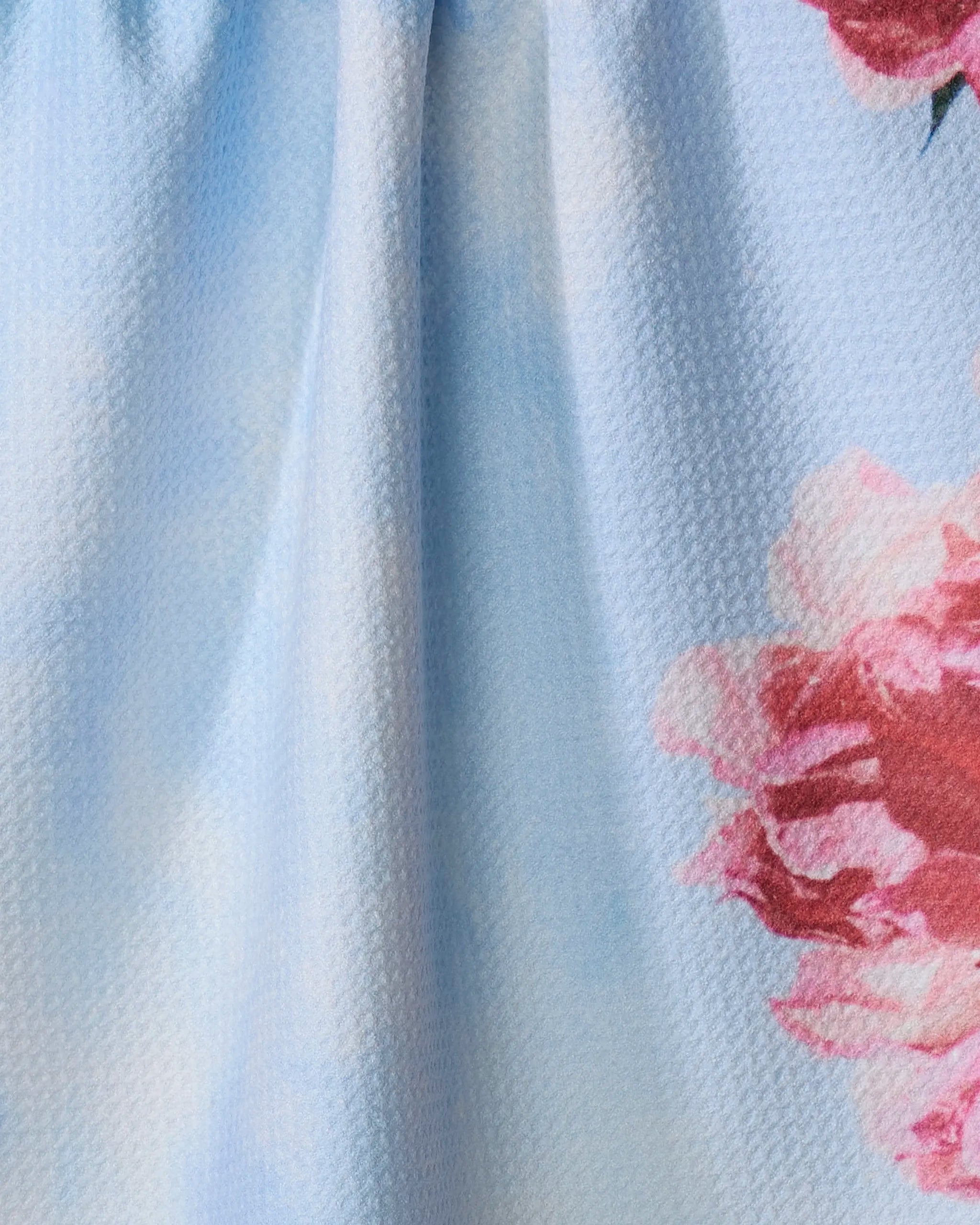 Close-up of the La Floraison kitchen towel by Stefanie Bales in light blue, showcasing textured fabric and large pink and white flowers on the right. The soft material is slightly gathered at the top for a charming touch.