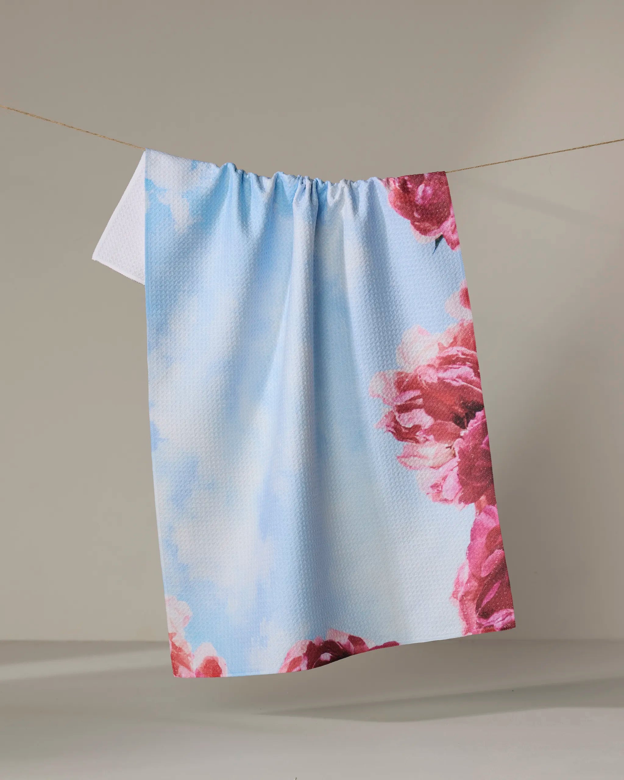 La Floraison by Stefanie Bales, a sky-blue tea towel with white clouds and pink peonies in the corners, made from recycled materials, hangs on a clothesline against a neutral wall.