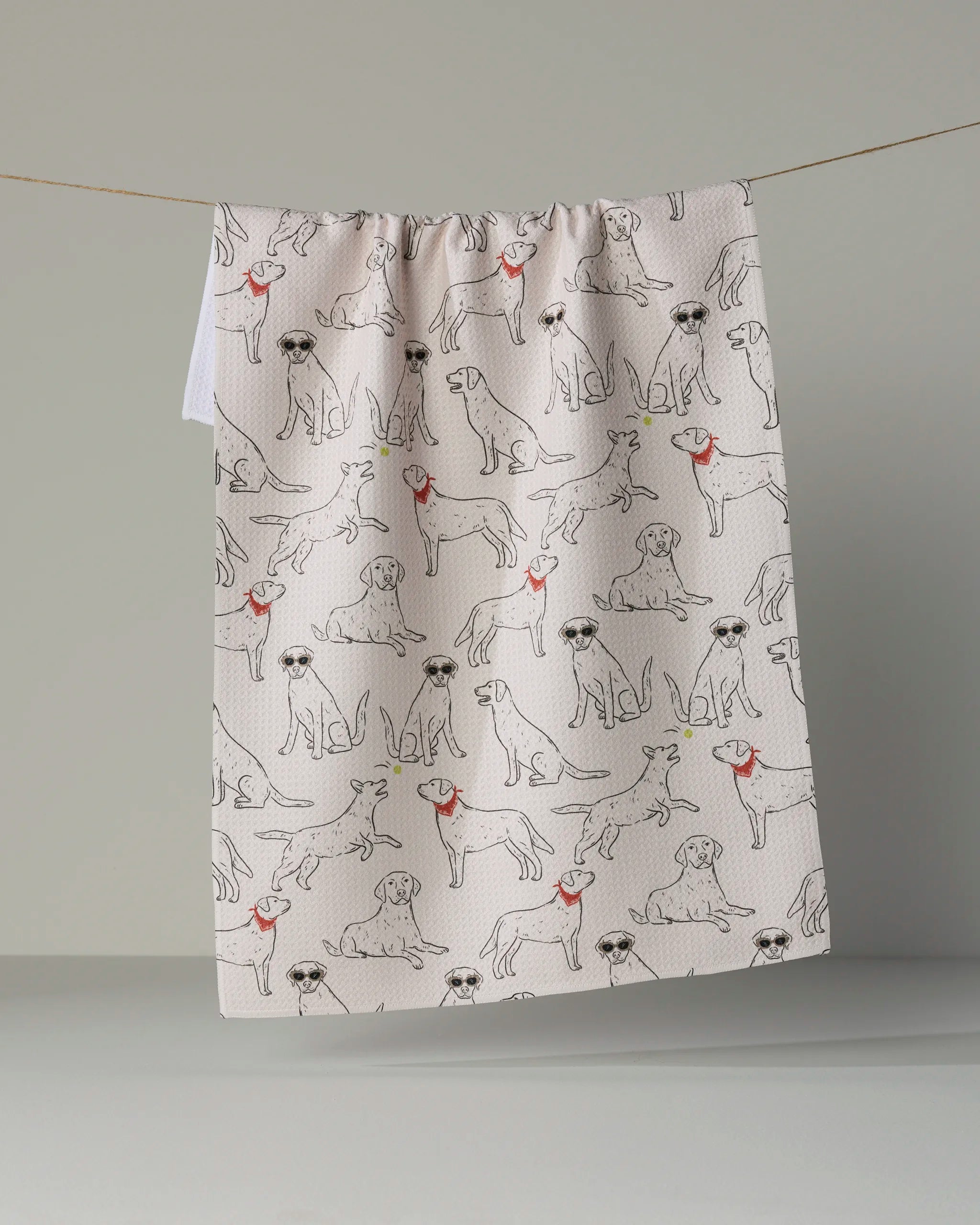 Add a playful touch to your kitchen décor with the “Lab Pack” tea towel by Geometry, featuring a charming pattern of illustrated dogs sporting sunglasses and red collars or bandanas on a light background—perfect for brightening up your space.