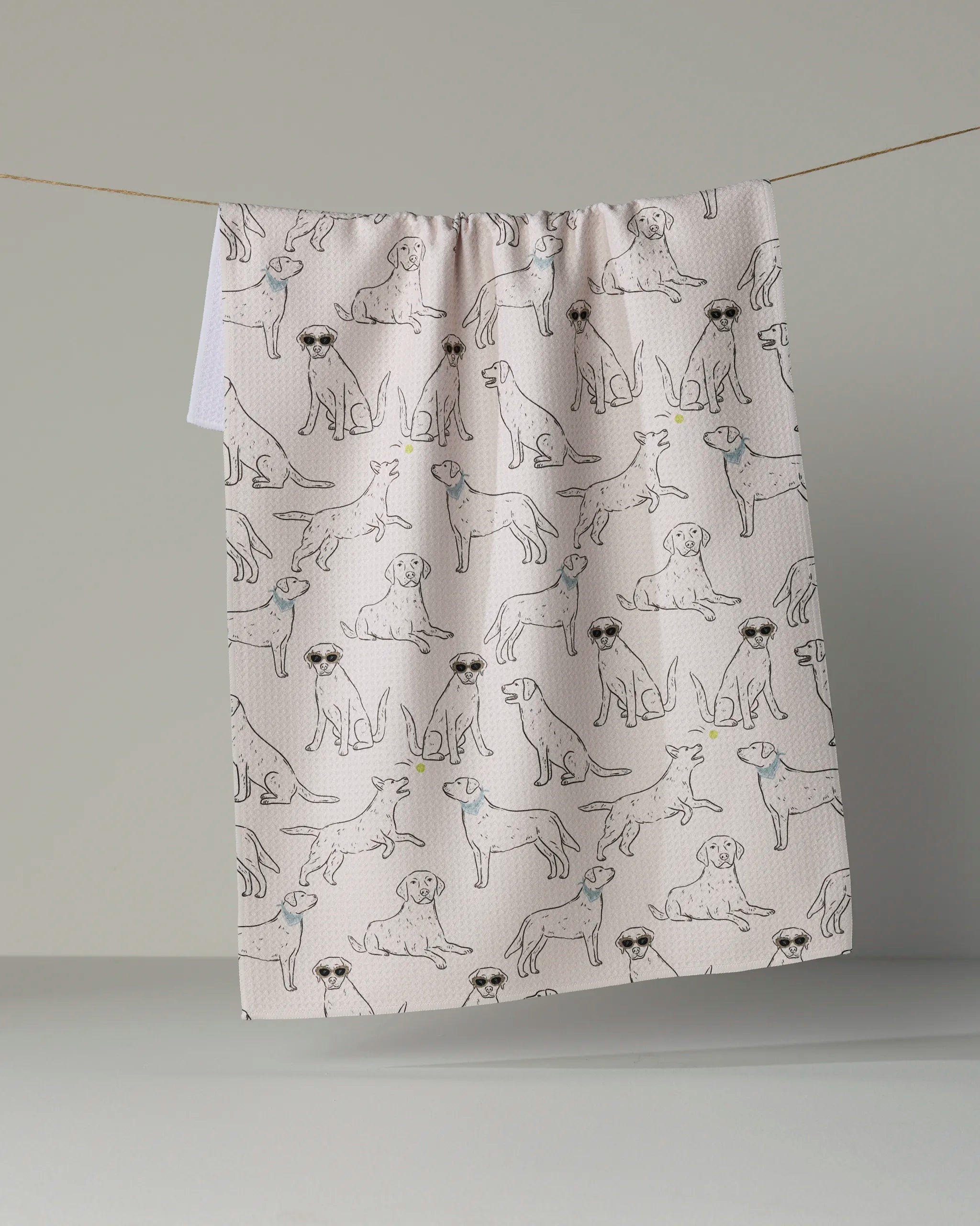 A white kitchen towel from Geometry’s Lab Pack collection, crafted from recycled materials and featuring a repeating pattern of outlined dogs wearing sunglasses, hangs on a line against a plain, neutral background.