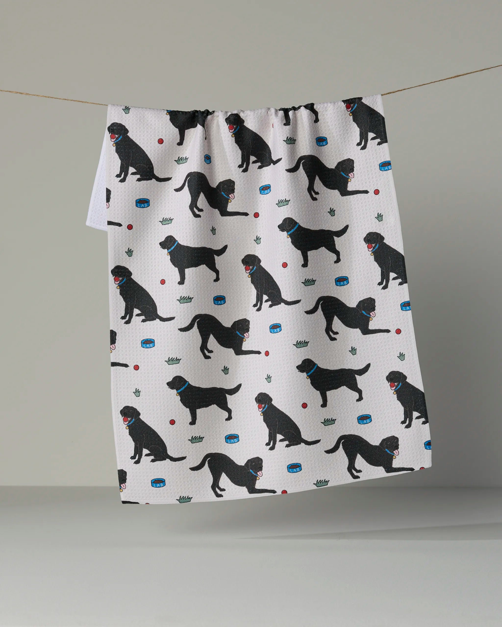 Geometry “Lab Pals” recycled kitchen towel featuring a repeating pattern of black dogs in various poses, along with paws, red balls, and green crowns, displayed hanging from a string against a plain background.