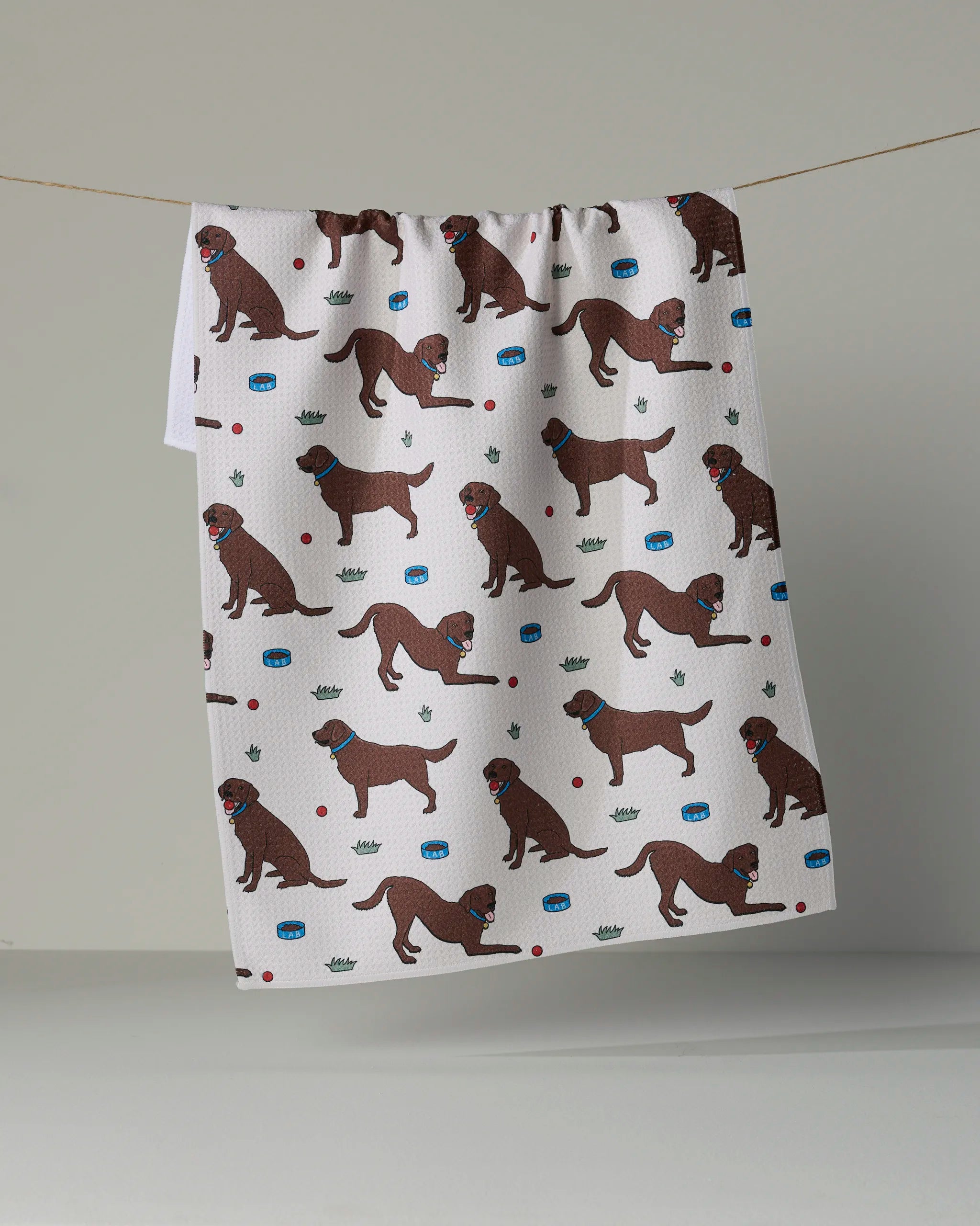 A white recycled kitchen towel from Geometry, called “Lab Pals,” hangs on a line, featuring a repeating pattern of brown dogs wearing blue collars, along with tennis balls, dog bowls, and green crowns on a plain background.