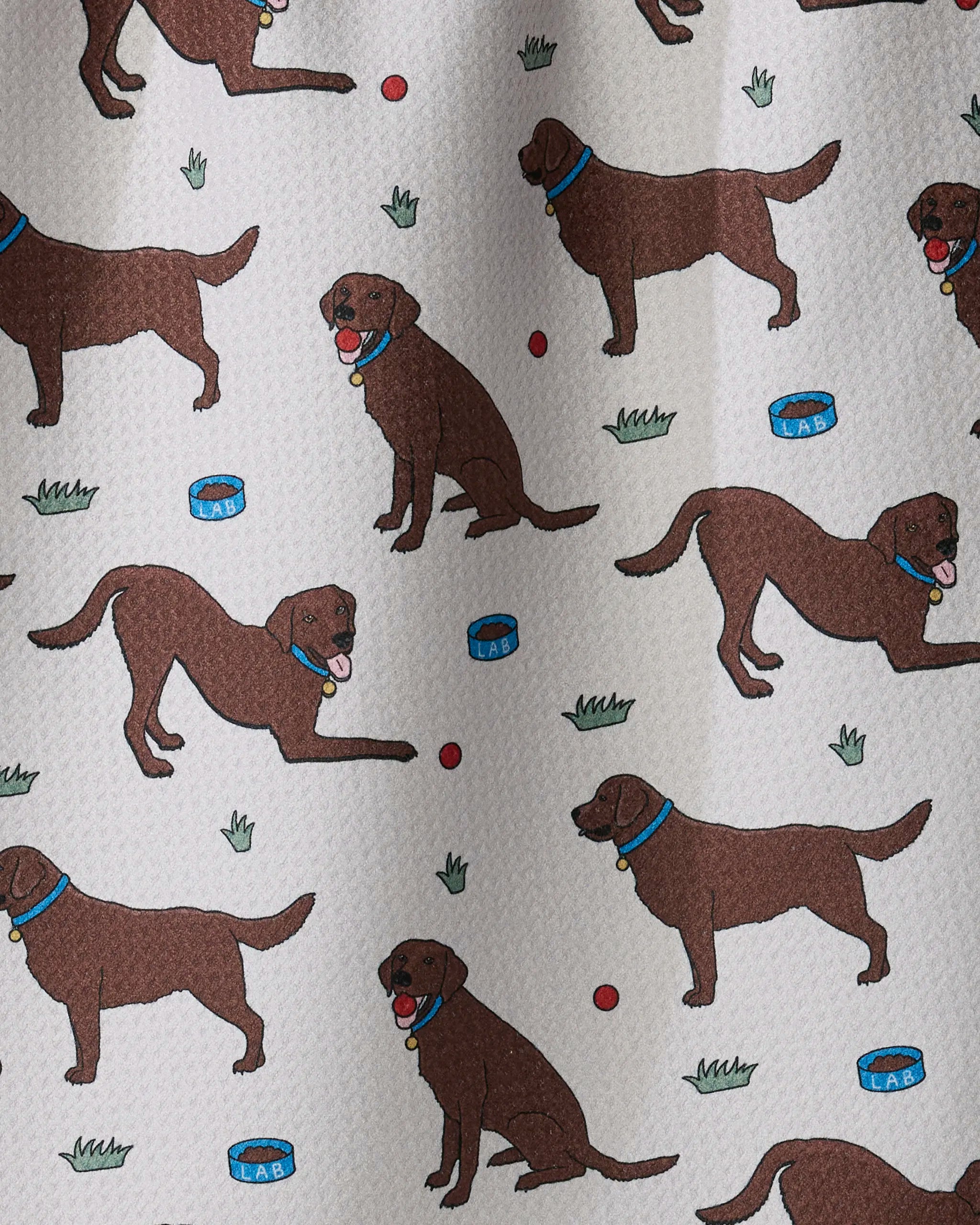 Pattern of cartoon brown dogs with blue collars in various poses, red balls, blue dog bowls labeled “LAB,” and green tufts of grass on a light background—the Lab Pals recycled kitchen towel by Geometry is ultra-absorbent and perfect for your kitchen.