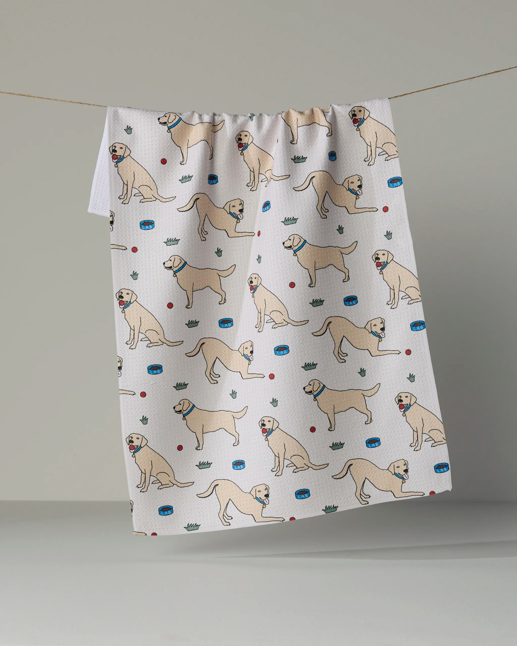 A “Lab Pals” tea towel by Geometry hangs on a line, featuring playful illustrations of yellow dogs in various poses, along with red balls, green crowns, and blue dog bowls.