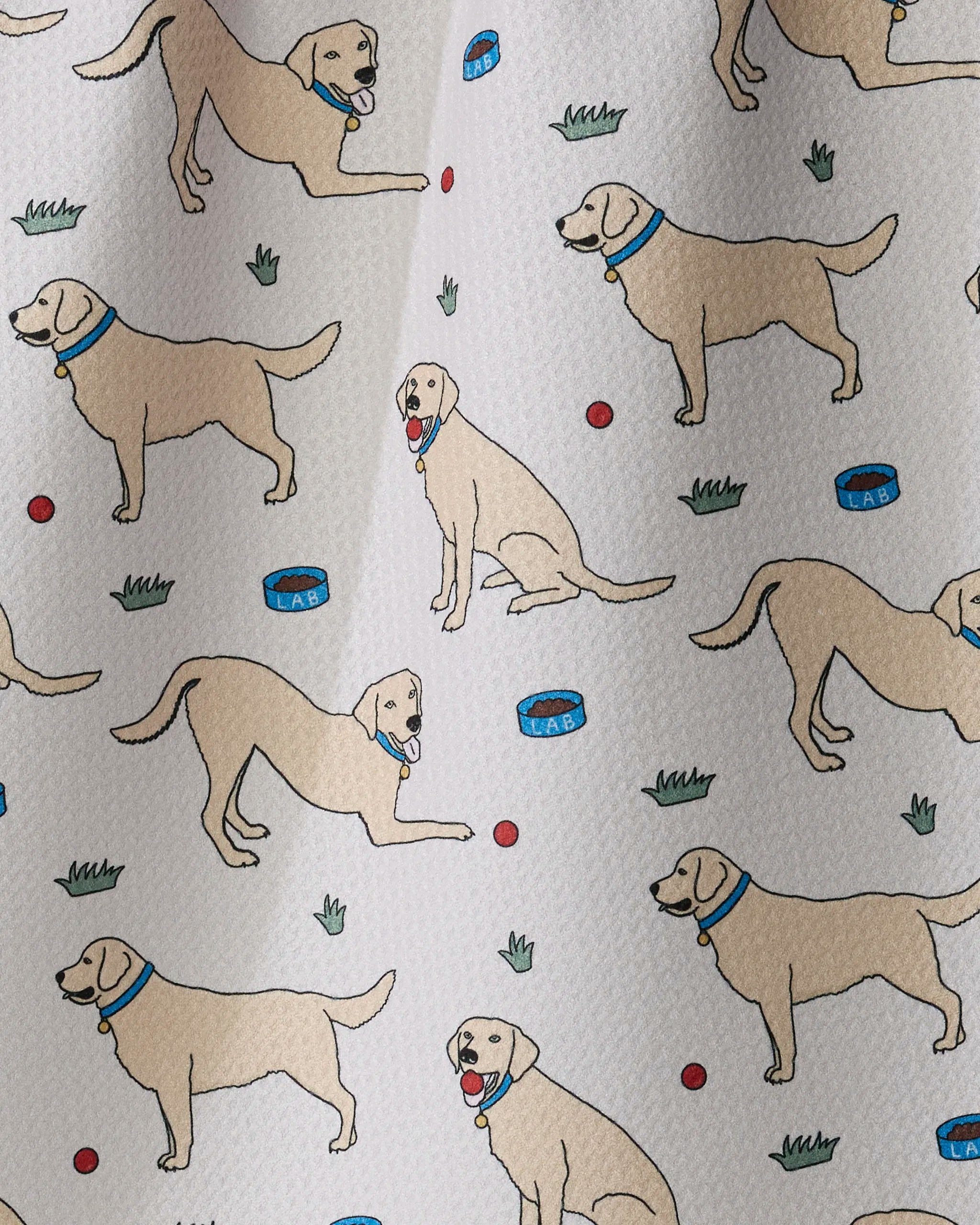 Geometry’s Lab Pals fabric showcases illustrated yellow Labradors with blue collars, red balls, blue bowls, and grassy accents on a white textured background—making it the perfect choice for an ultra-absorbent tea towel that brings cheerful charm to any kitchen.