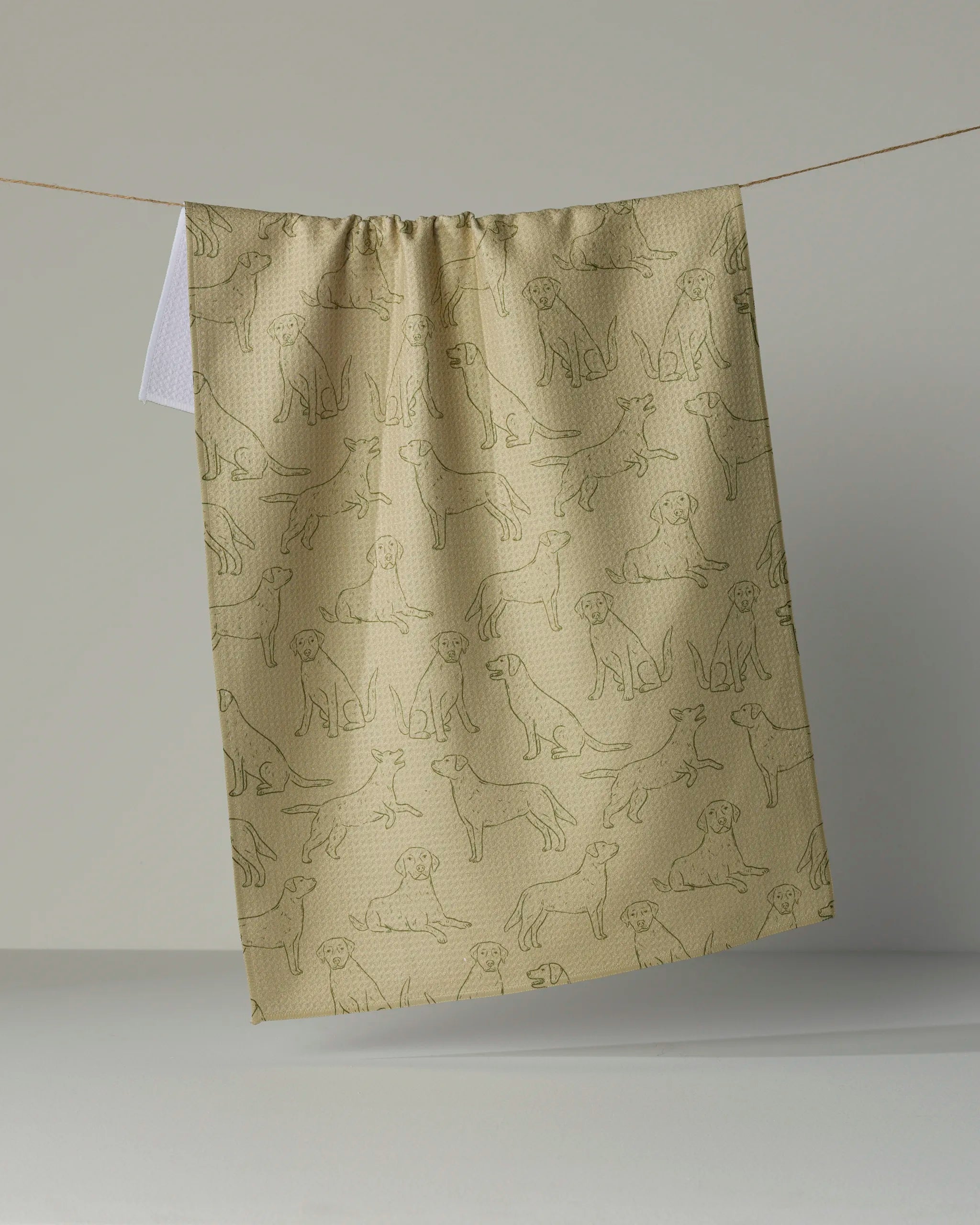 A tea towel from Geometry, called “Lab Pups,” featuring a tan background and simple outlined dog illustrations, hangs from a string against a neutral backdrop. It’s crafted from ultra-absorbent recycled materials.