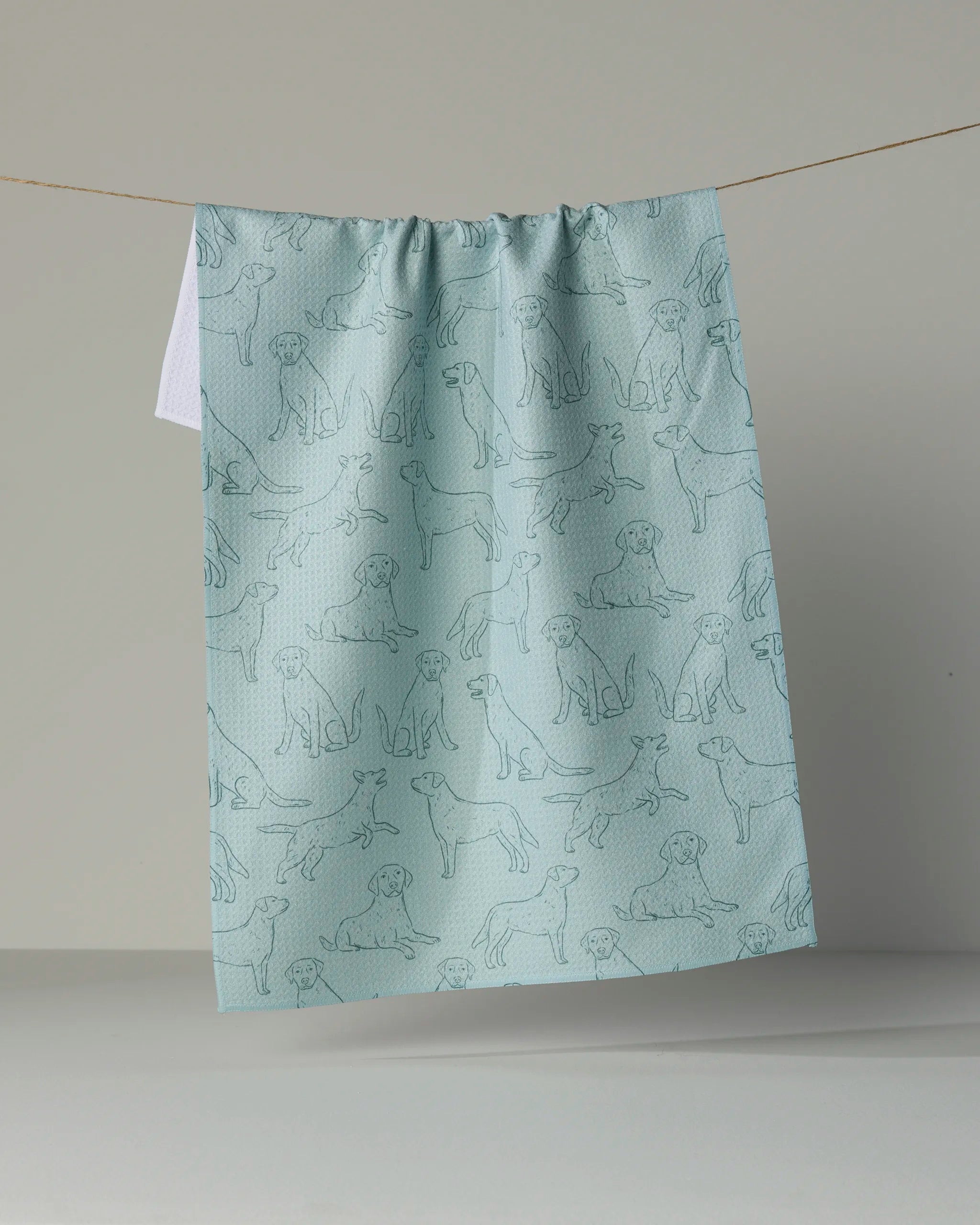 A light blue, ultra-absorbent “Lab Pups” tea towel by Geometry, featuring line drawings of various dogs in different poses, crafted from recycled materials and hanging from a string against a plain background.