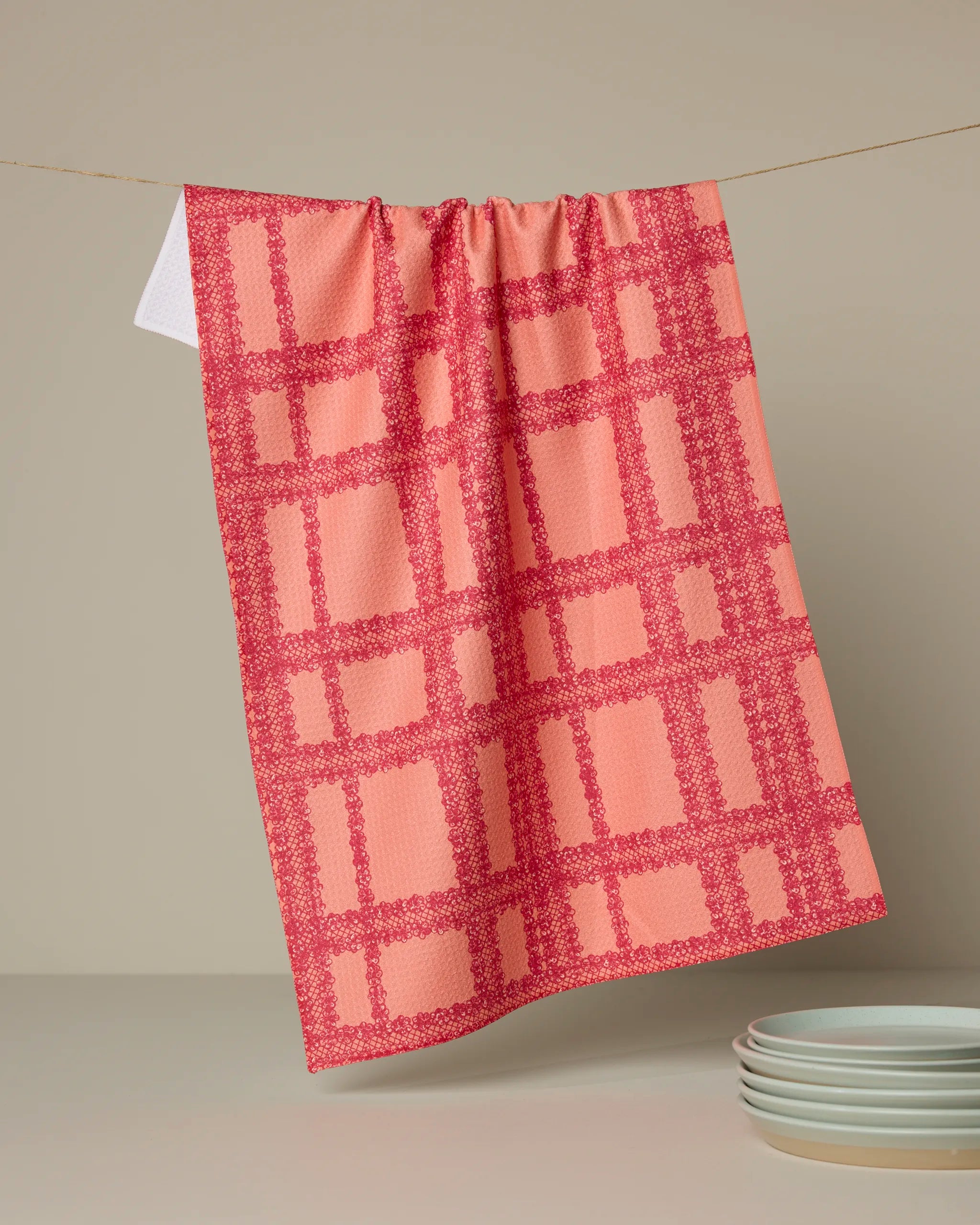 The Lacy Plaid tea towel by Geometry, featuring pink and red checks, hangs on a line against a beige background with white plates below. This quick-drying towel brings charm and function to any kitchen.