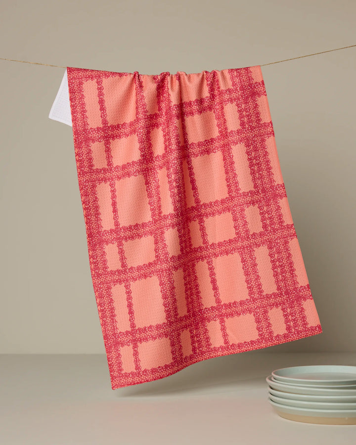 The Lacy Plaid tea towel by Geometry, featuring pink and red checks, hangs on a line against a beige background with white plates below. This quick-drying towel brings charm and function to any kitchen.
