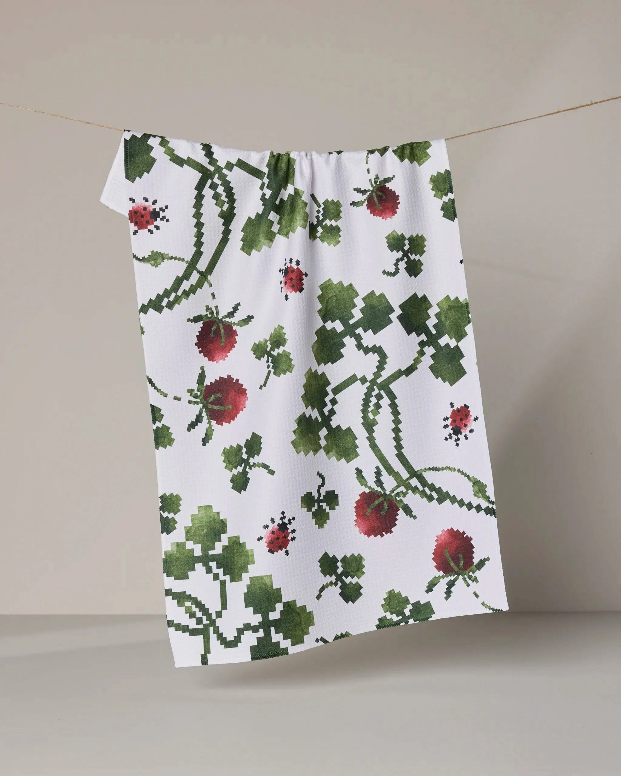 Geometry’s “Ladybug Fields” tea towel, featuring a pixelated red strawberry, green leaf, and ladybug pattern, hangs against a plain light gray background.