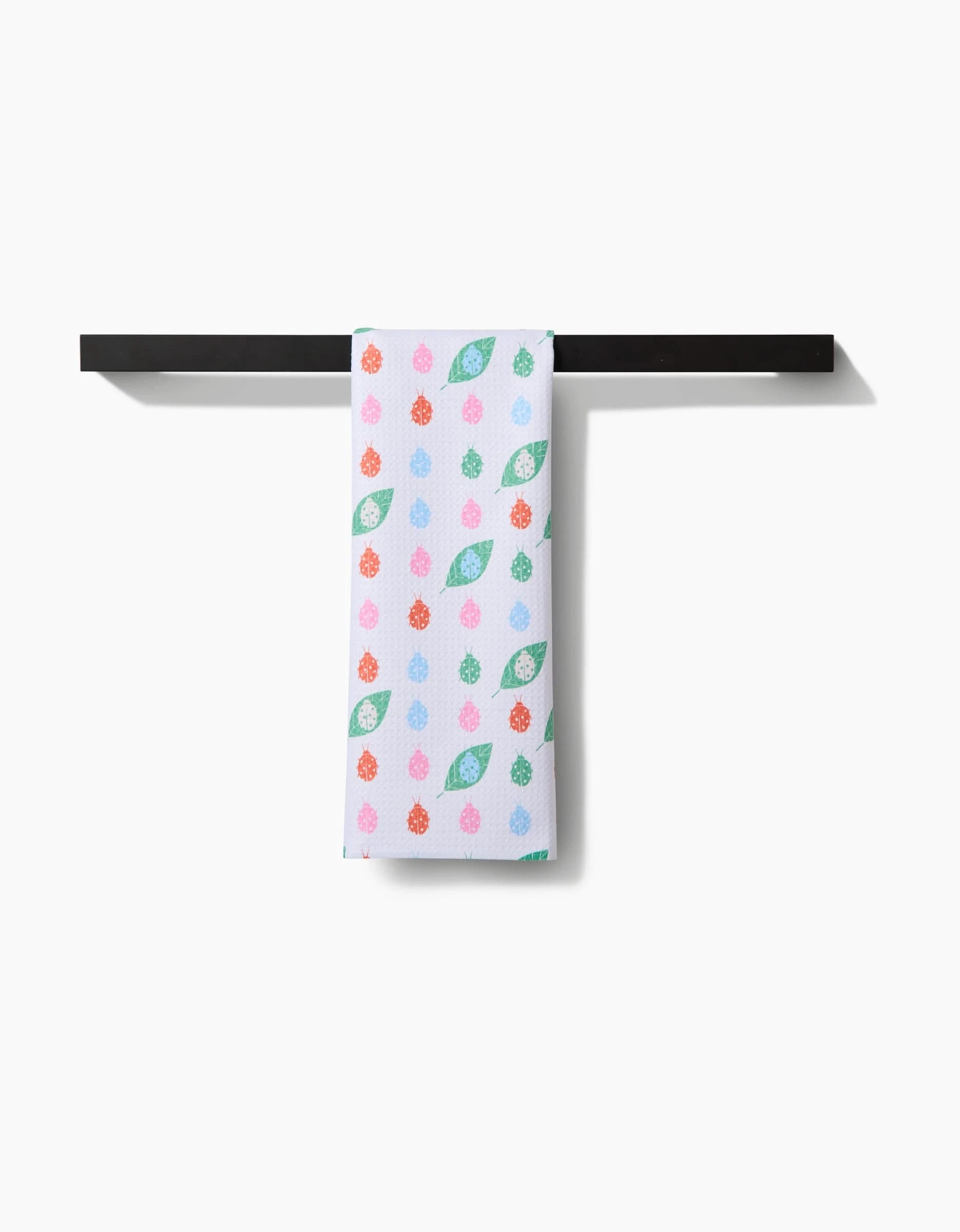 The Ladybug Leaf tea towel by Elisabeth Wing, crafted from recycled materials and adorned with green leaves and colorful berries, hangs neatly on a black towel bar against a plain white background.