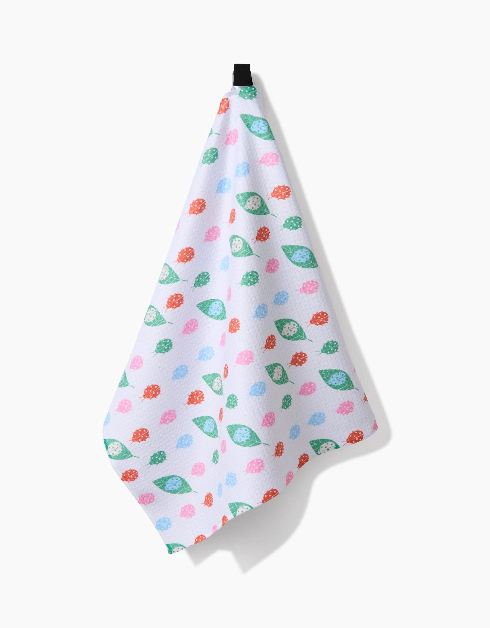 The Ladybug Leaf tea towel by Elisabeth Wing is white, made from recycled materials, and features a vibrant pattern of strawberries and green leaves. It hangs from a black hook against a plain white background.