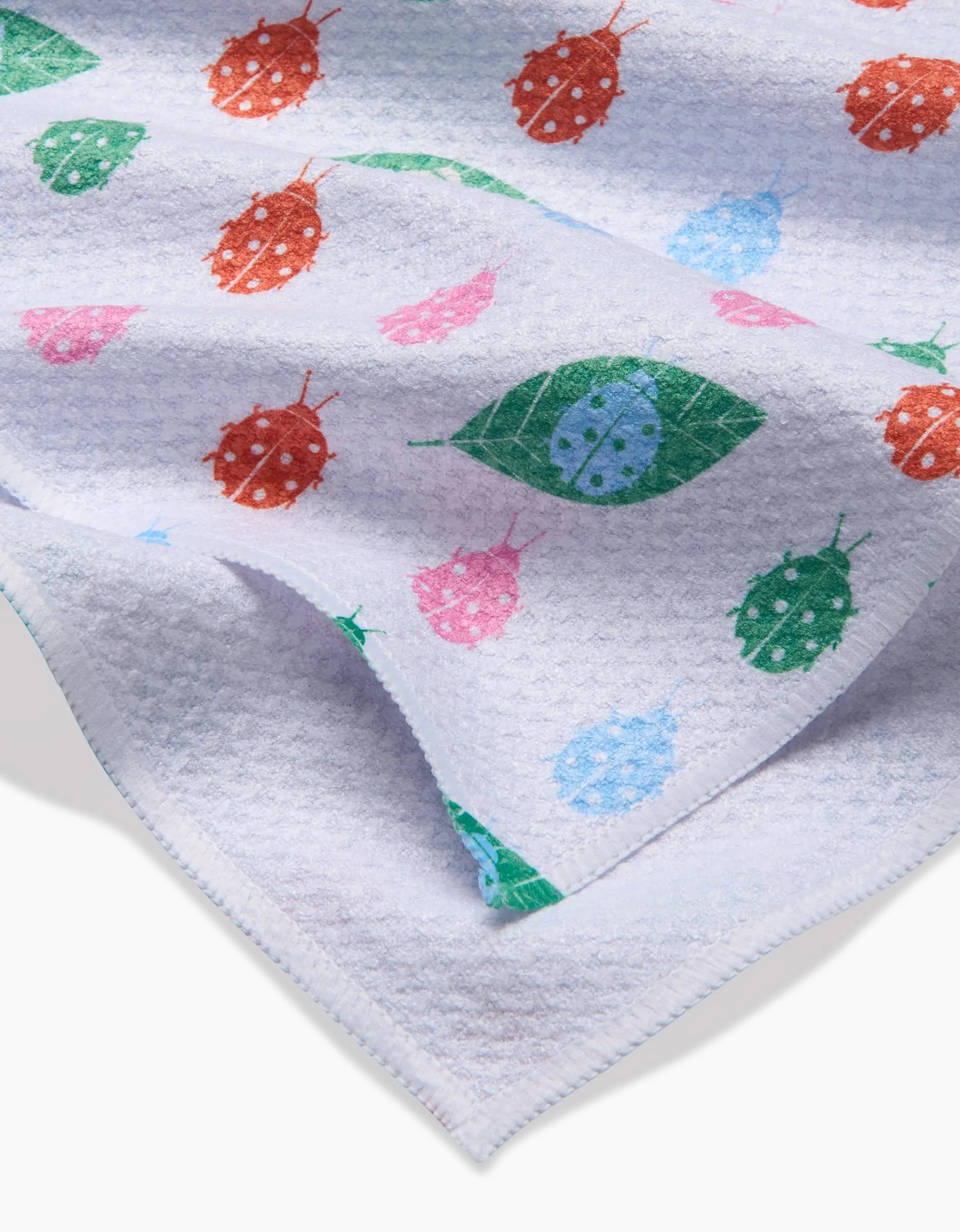 The Ladybug Leaf tea towel by Elisabeth Wing features colorful ladybugs and green leaves on white Geoweave fabric, with a soft, lightweight texture—perfect for adding eco-friendly charm to your kitchen.
