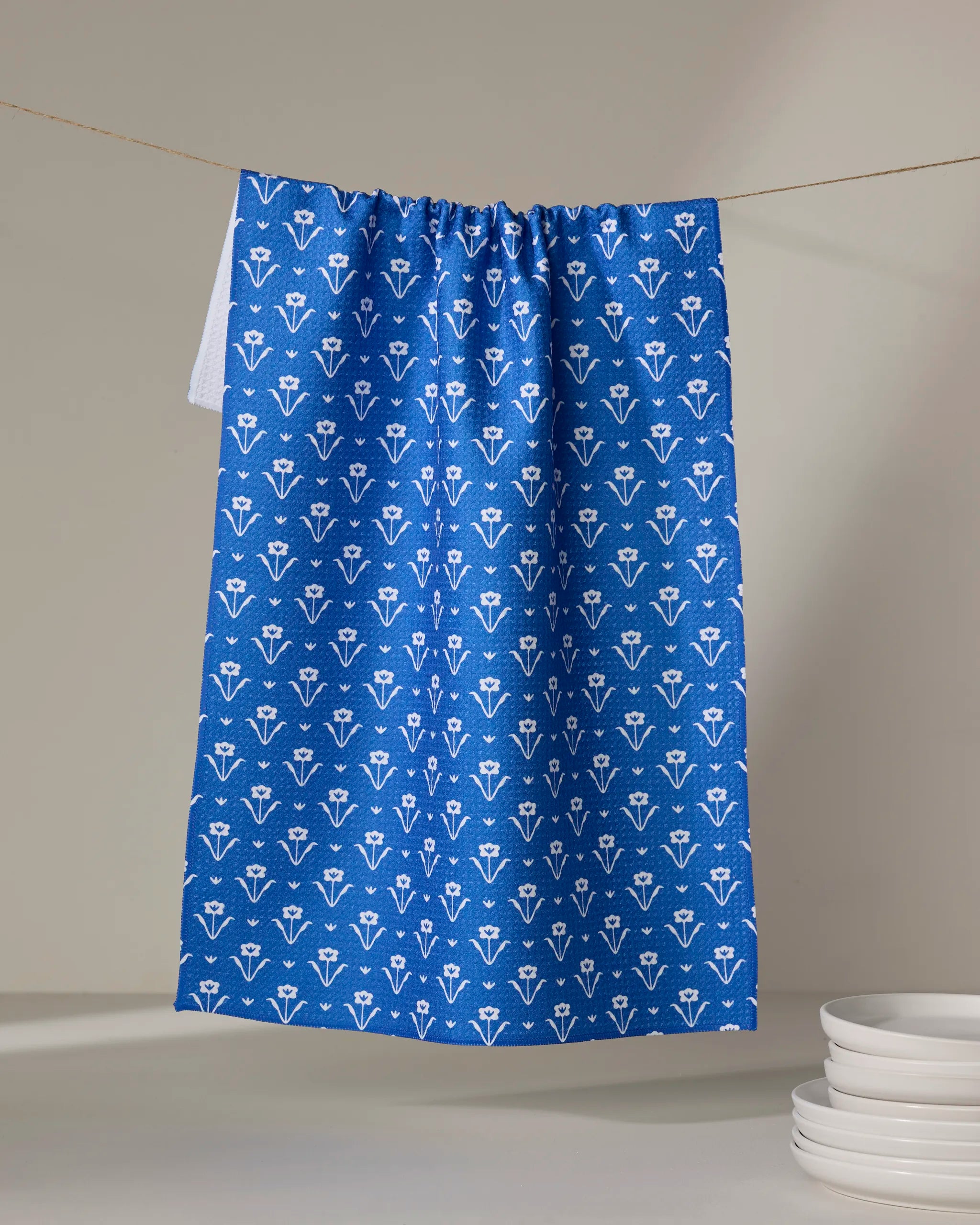 The Buy Guide's Ladylake - Navy tea towel, featuring a white floral pattern, hangs on a clothesline above a small stack of white plates on the right below.