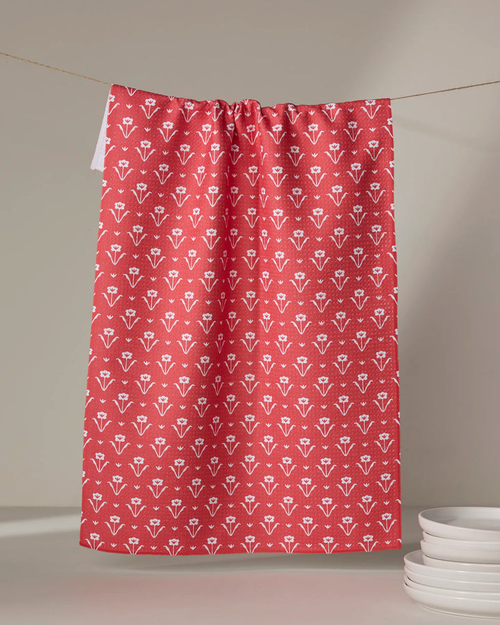 A Ladylake - Red kitchen tea towel by The Buy Guide, featuring a white floral pattern, hangs on a string. Nearby, a small stack of white plates rests on a light surface.