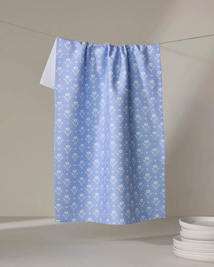 The Ladylike - French Blue tea towel by The Buy Guide, featuring a white floral pattern and a hint of white at the top left, hangs on a line with four stacked white plates in the bottom right on a light gray background.