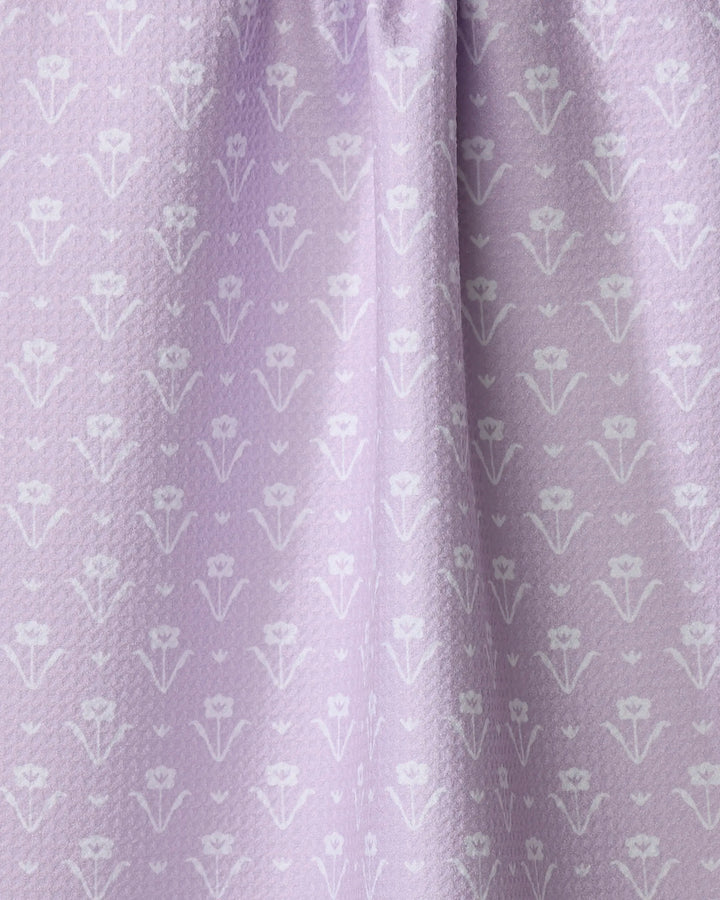 A close-up of Ladylike - Lilac by The Buy Guide shows light purple fabric with a subtle textured weave and a repeating pattern of small, stylized white tulip-like flowers, softly gathered at the top—ideal for tea towels.