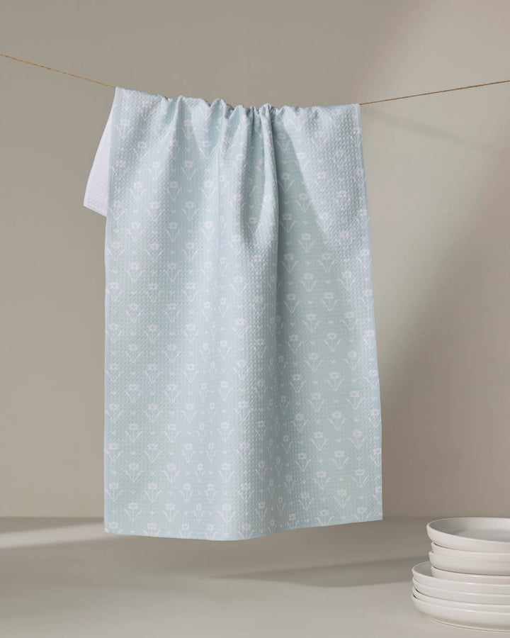 The Buy Guide’s Ladylike - Mint dish towel, featuring a light blue pattern, hangs on a clothesline against a neutral background, with white plates stacked below.