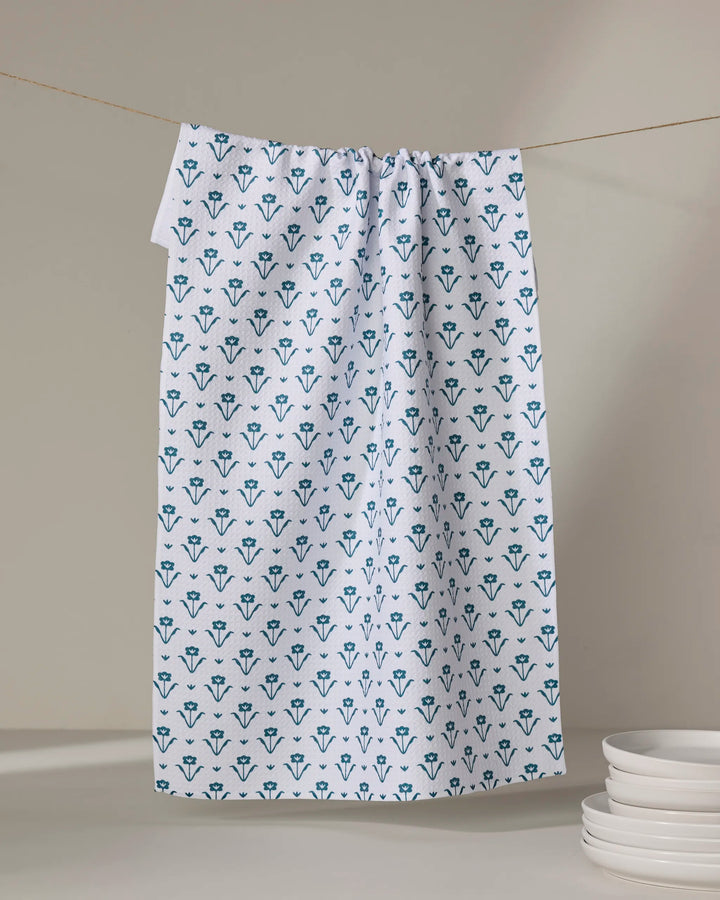 The Buy Guide's Ladylike Too - Emerald, an eco-friendly kitchen towel with a blue floral pattern, hangs on a clothesline beside a stack of white plates on a beige surface.