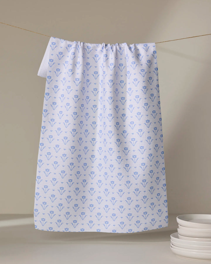 The Ladylike Too - French Blue tea towel by The Buy Guide, featuring a repeating blue floral pattern, hangs above a stack of white plates against a neutral background for a fresh, inviting kitchen display.