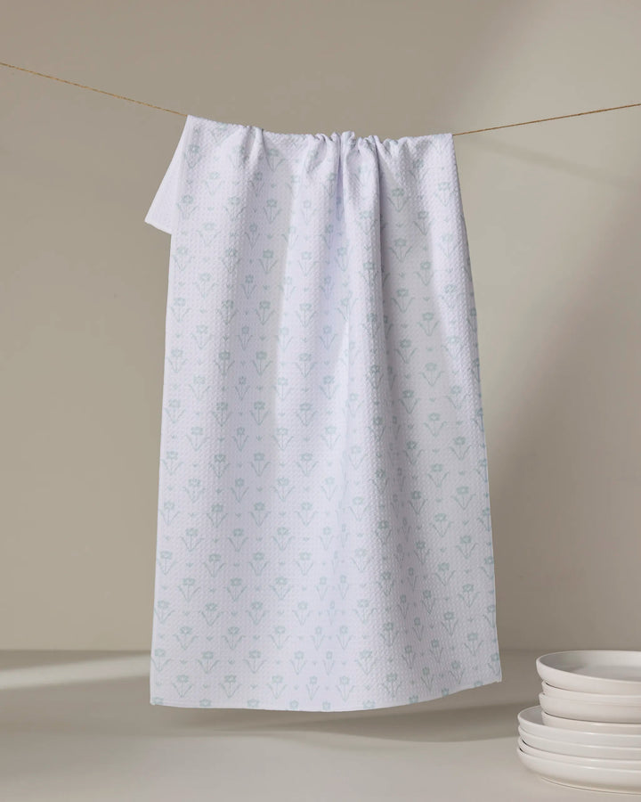The Buy Guide's Ladylike Too - Mint tea towel, featuring a subtle light blue floral pattern, hangs from a string against a beige background above a stack of white plates.