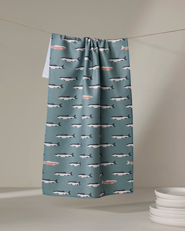 The Lake Fish tea towel by Coit Creative, made from recycled materials, features teal fabric with swimming whales and pink fish. Ultra-absorbent, it's pictured hanging on a line near stacked white plates on a light surface.