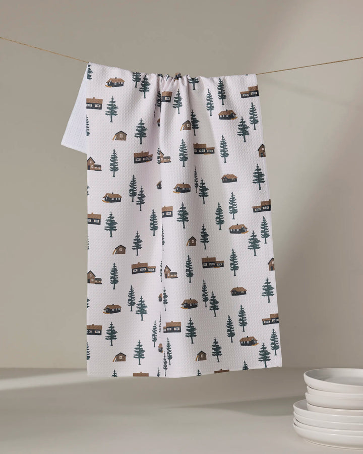 The Coit Creative Lakehouse Cabins tea towel, made from recycled materials, displays a cabins and trees pattern. It hangs on a clothesline above stacked plates, set against a simple neutral background.