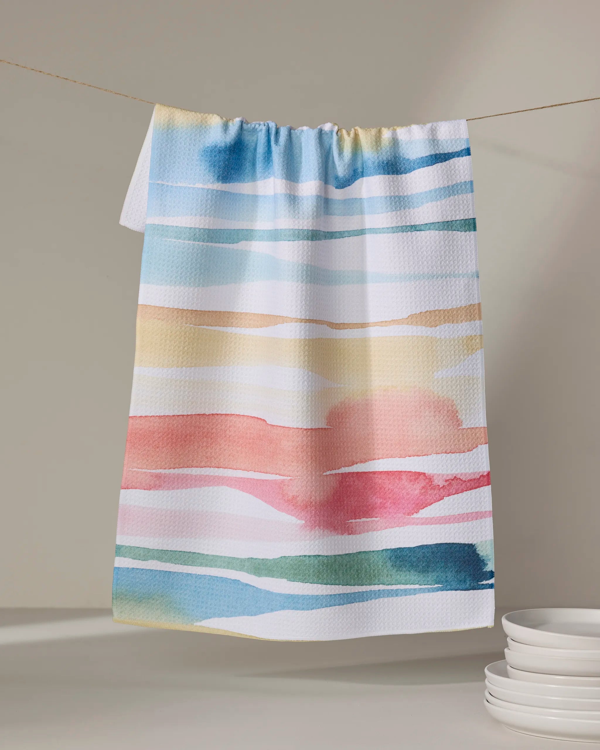 The “Lakeside” tea towel by Sara Mari, featuring horizontal watercolor stripes in blue, yellow, peach, and green, hangs above a stack of white plates against a neutral background.