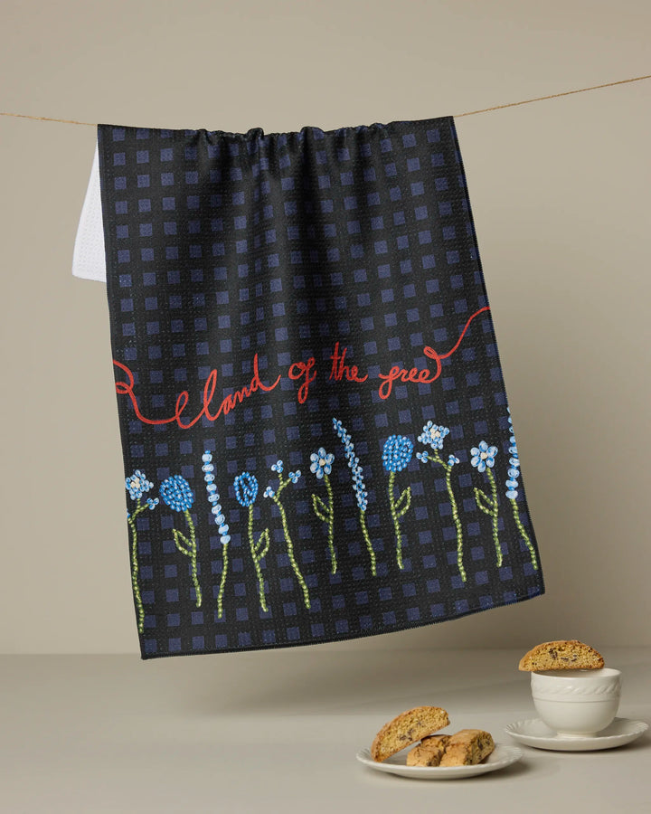 The Geometry "Land of Free" dark checkered tea towel, featuring embroidered blue flowers and “land of the free” in red script, hangs above a table set with biscotti on a plate and in a bowl.