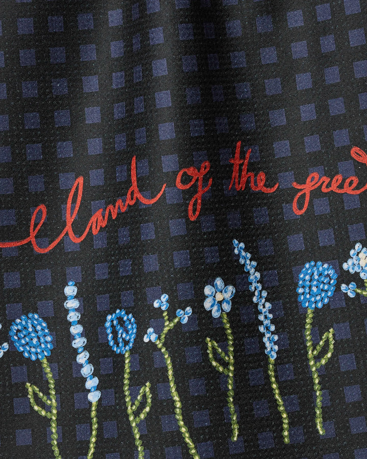 A close-up of Geometry’s Land of Free tea towel, featuring a blue checkered pattern with embroidered blue and white flowers, green stems, and red cursive text that says “land of the free.”.