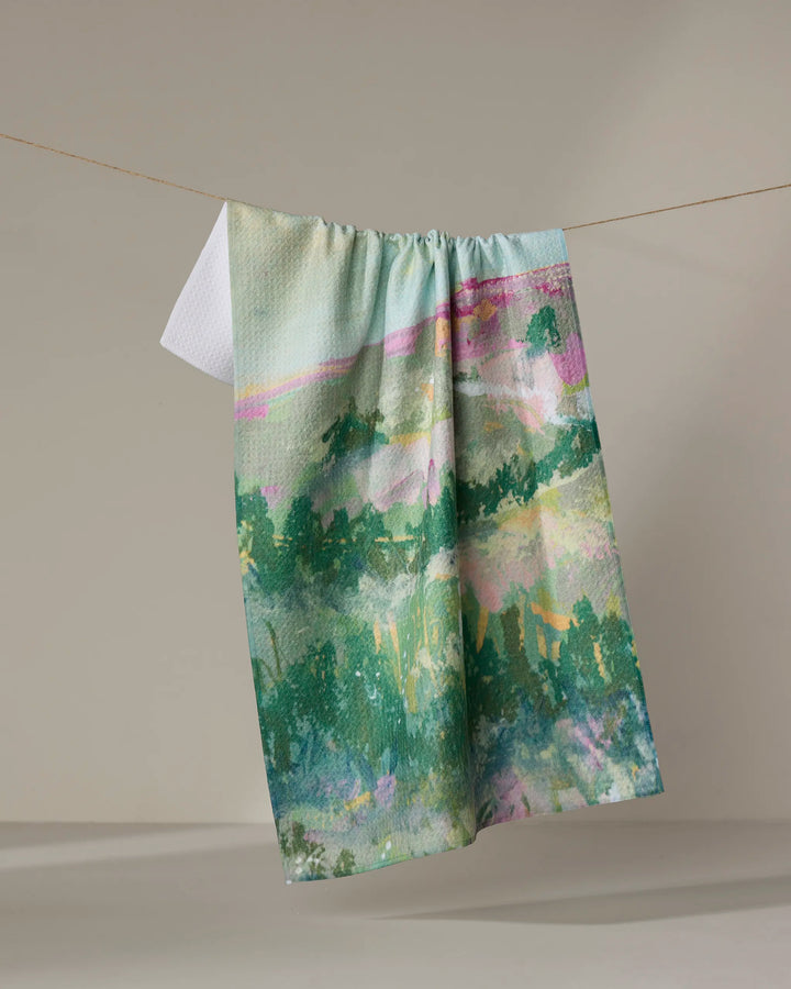 The "Land of Milk and Honey" recycled kitchen towel by Taryn Brennan, featuring an abstract watercolor landscape in green, blue, and pink, hangs on a string against a neutral background.