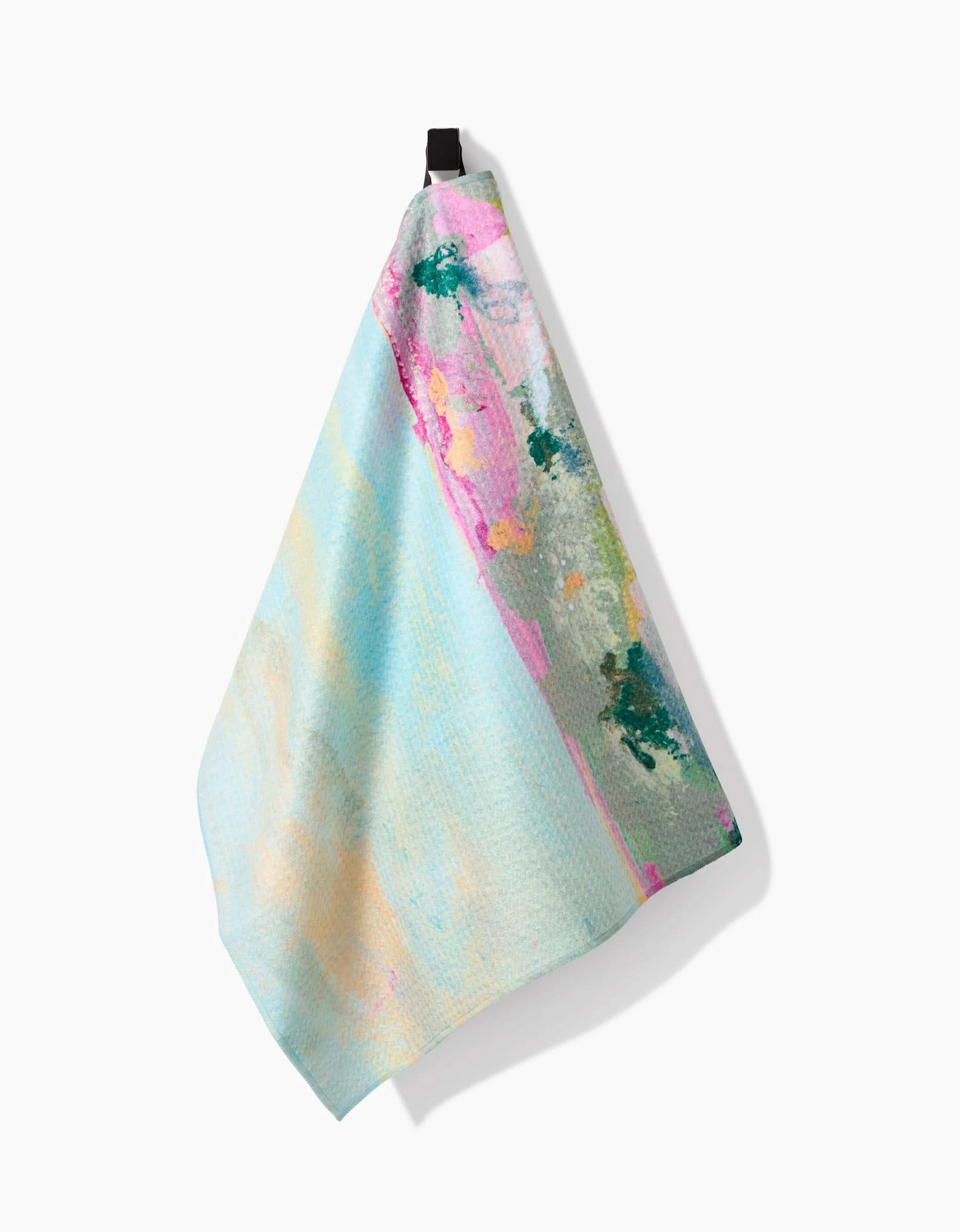 The Land of Milk and Honey tea towel by Taryn Brennan features pastel abstract patterns in blue, pink, yellow, and green on Geoweave fabric, hanging from a black loop against a white background.