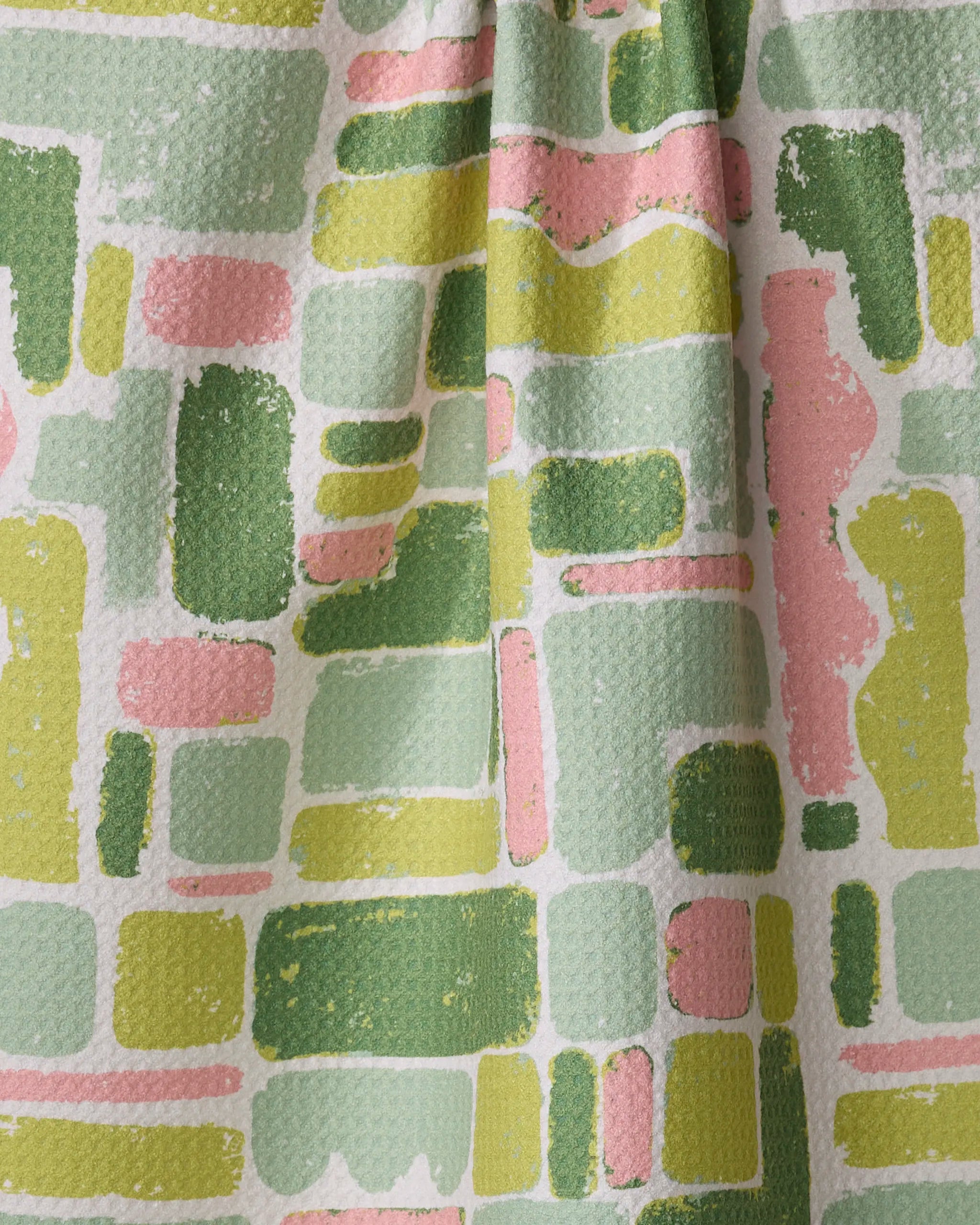 The Kayla Ann Landpatch is a textured, absorbent kitchen towel with an abstract pattern of overlapping rectangles and rounded shapes in green, yellow, pink, and white. Its slightly gathered material forms soft folds and subtle shadows.