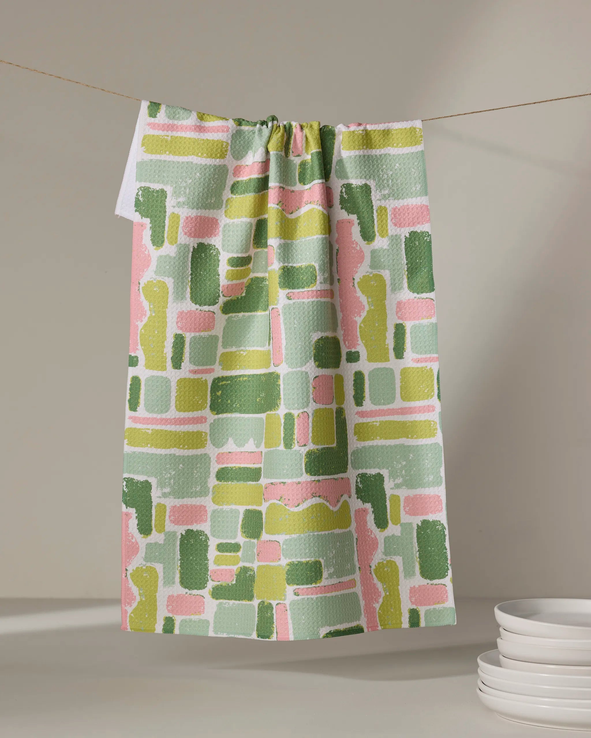 The Kayla Ann Landpatch kitchen towel, featuring an abstract geometric pattern in green, pink, and yellow, hangs on a clothesline near a stack of white plates against a neutral background.