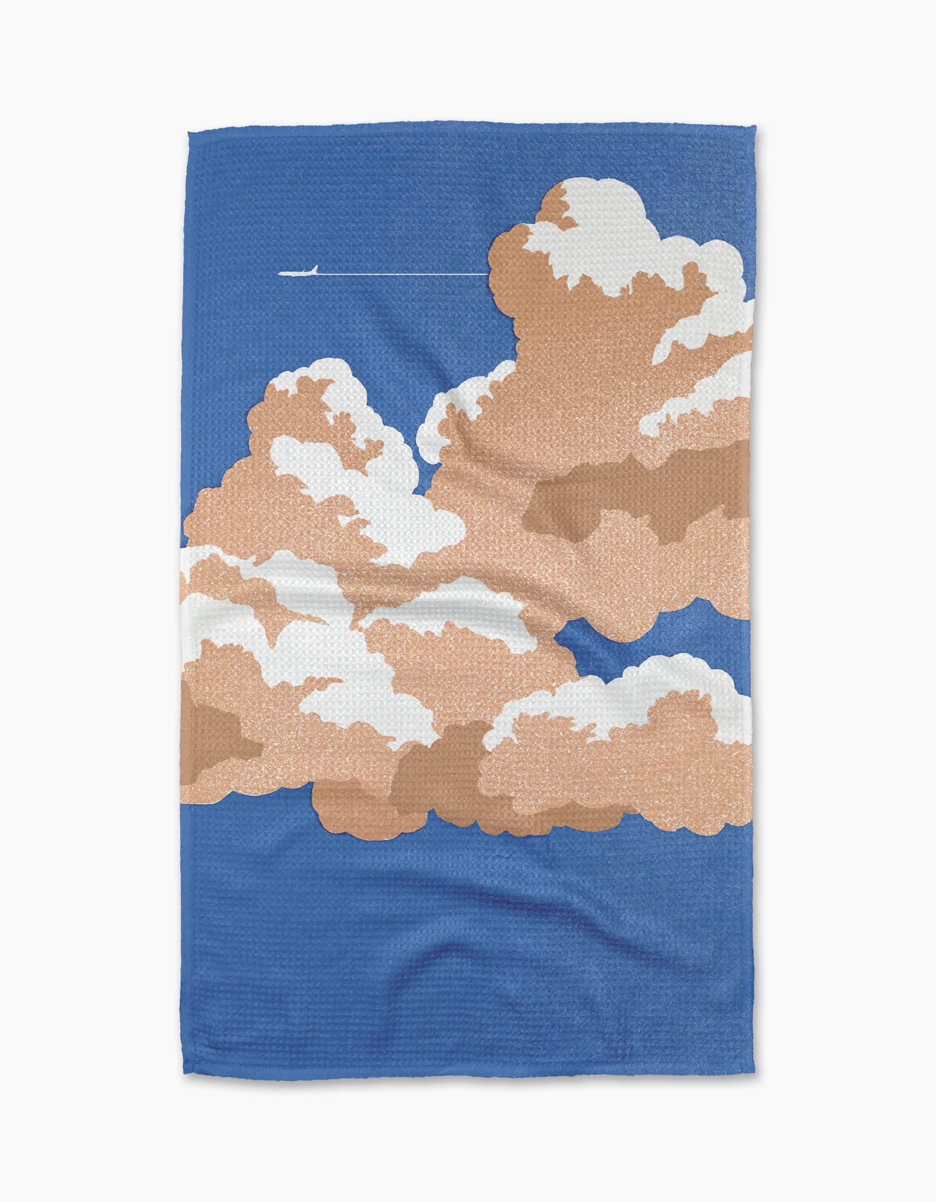 The Lani tea towel by Geometry features fluffy brown and white clouds with a small airplane and contrail on a blue sky, crafted from recycled materials.