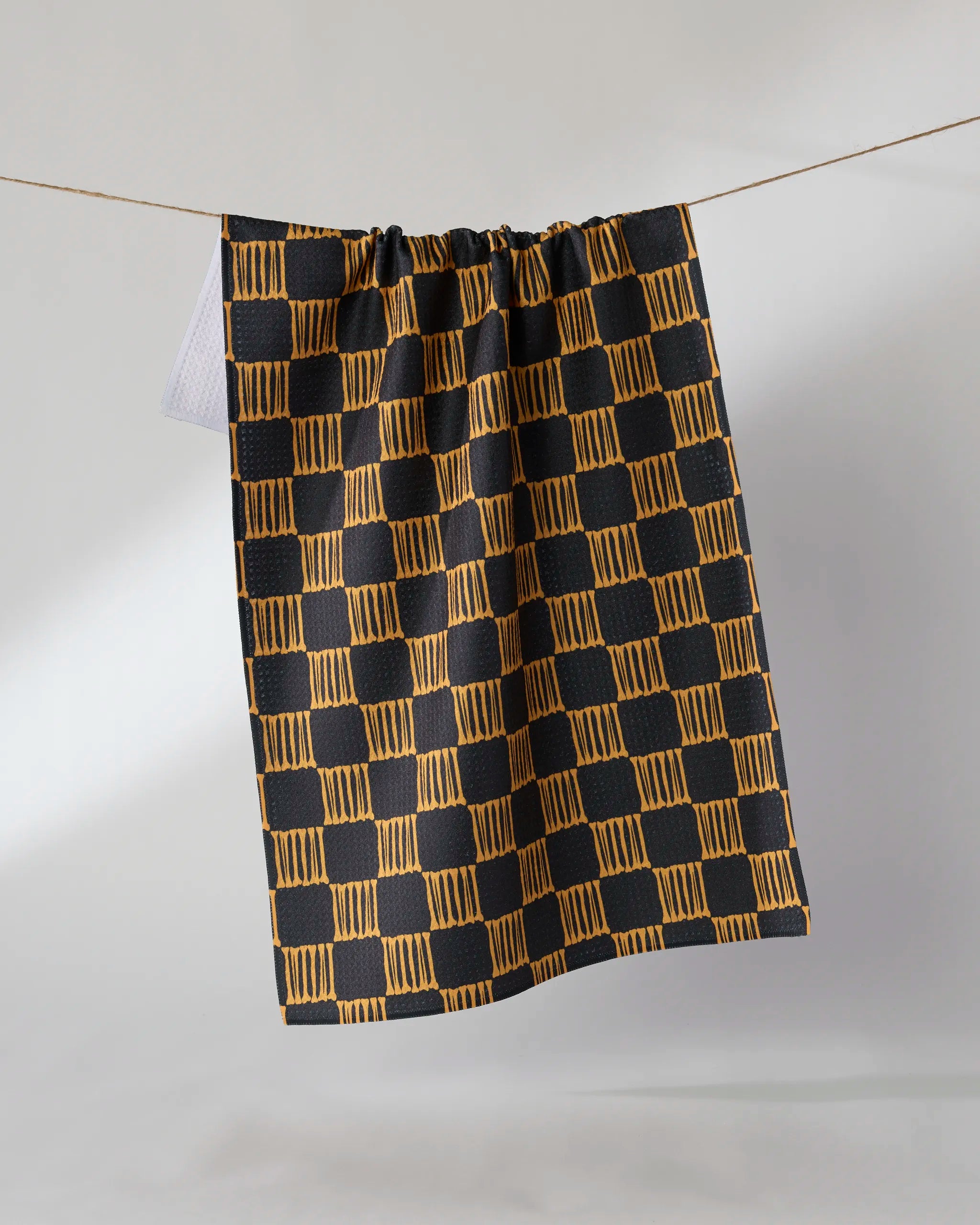 The Geometry Lantern Grid kitchen towel, featuring a black and gold checkered design and made from recycled materials, hangs on a clothesline against a plain light background.